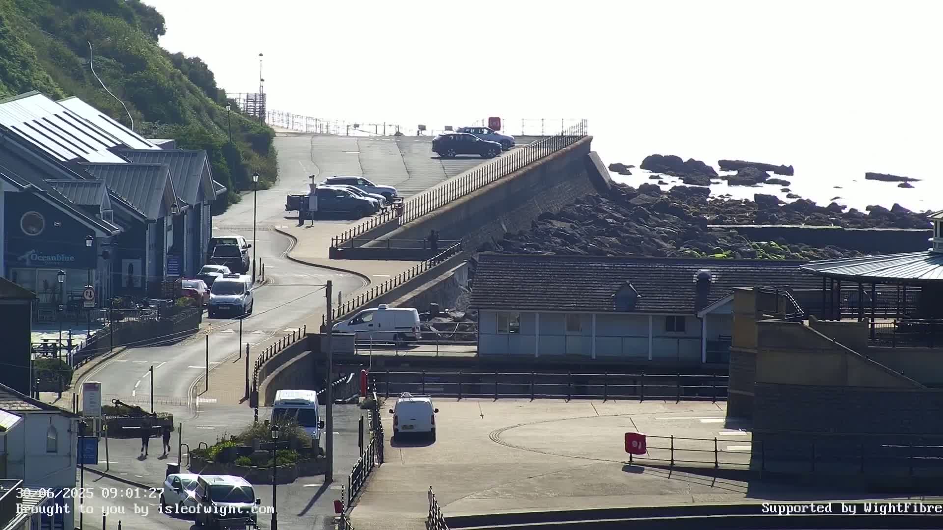A sunny seaside scene shows a coastal road with parked cars, buildings, and a seawall overlooking dark rocks and calm water.