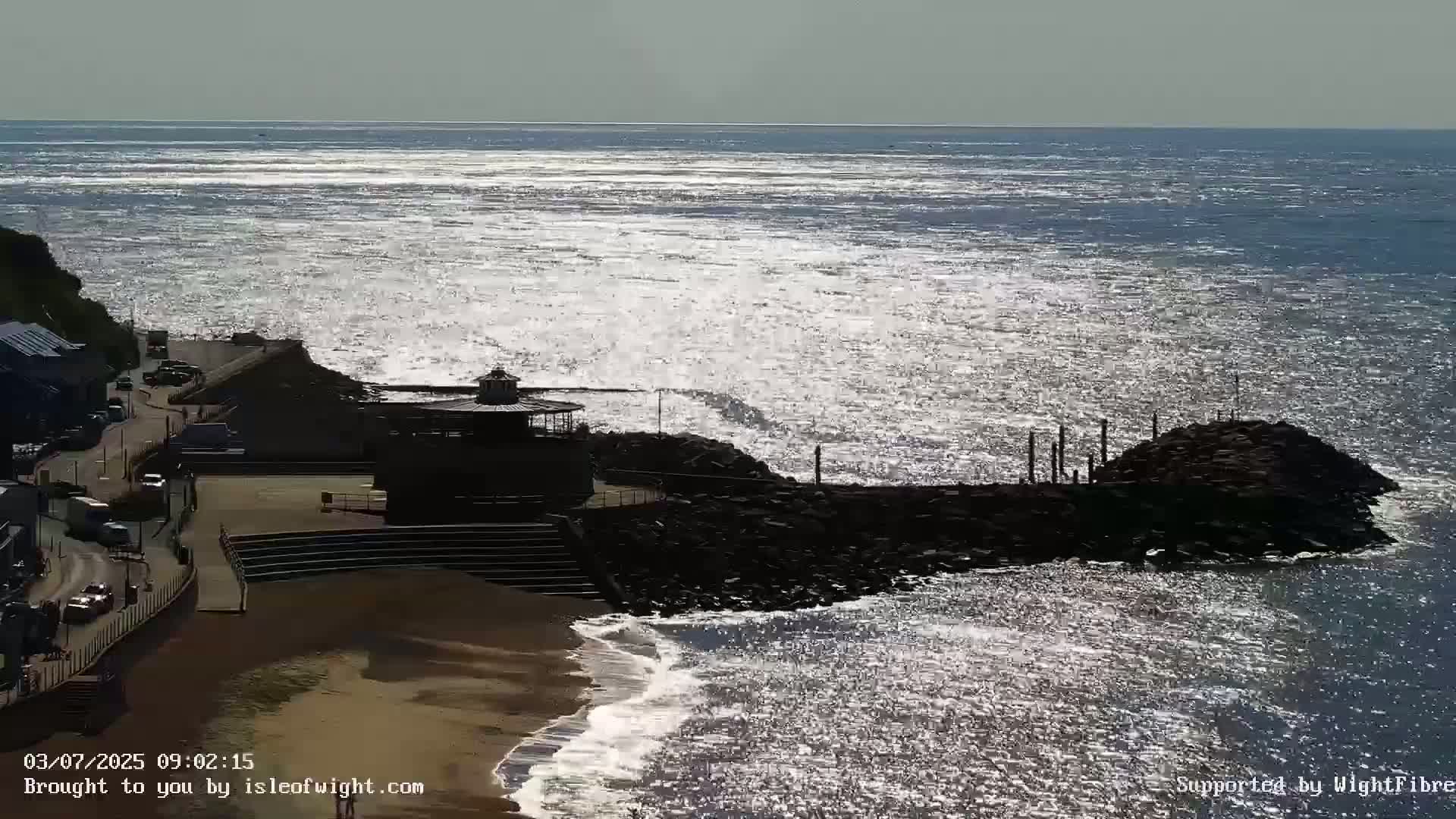 A sunlit beach scene shows a rocky outcropping, a small building, and a sandy beach next to a road with cars, all overlooking a sparkling ocean under a mostly cloudy sky.