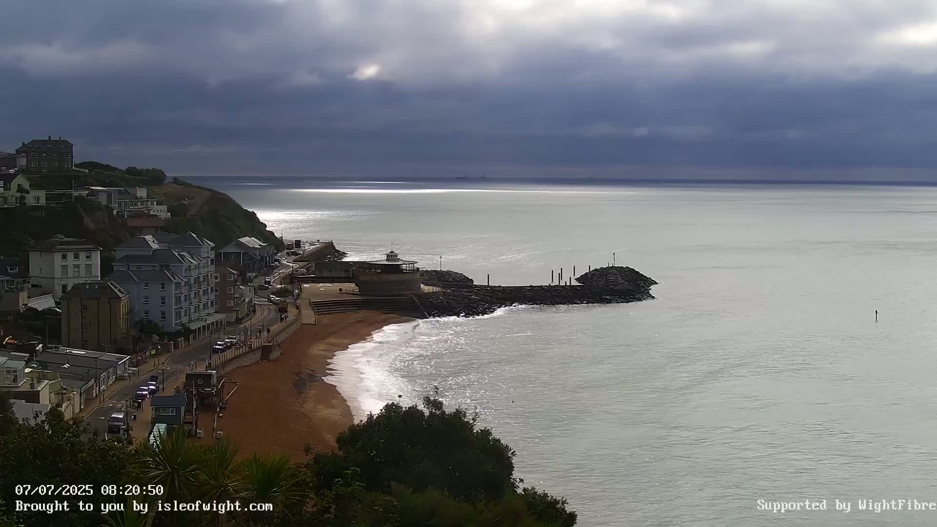 A coastal town with buildings on a cliff overlooking a sandy beach with gentle waves under a cloudy sky.