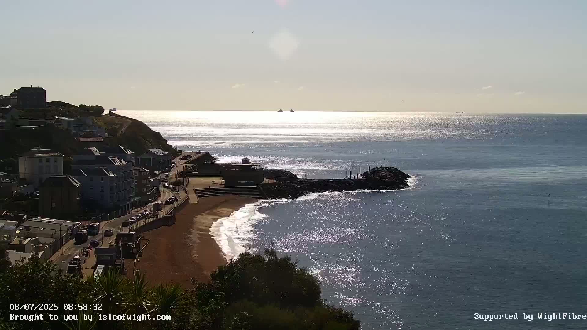 A sunny day at a seaside town shows a sandy beach with small waves lapping the shore, buildings on a cliff overlooking the calm ocean with a few ships on the horizon.