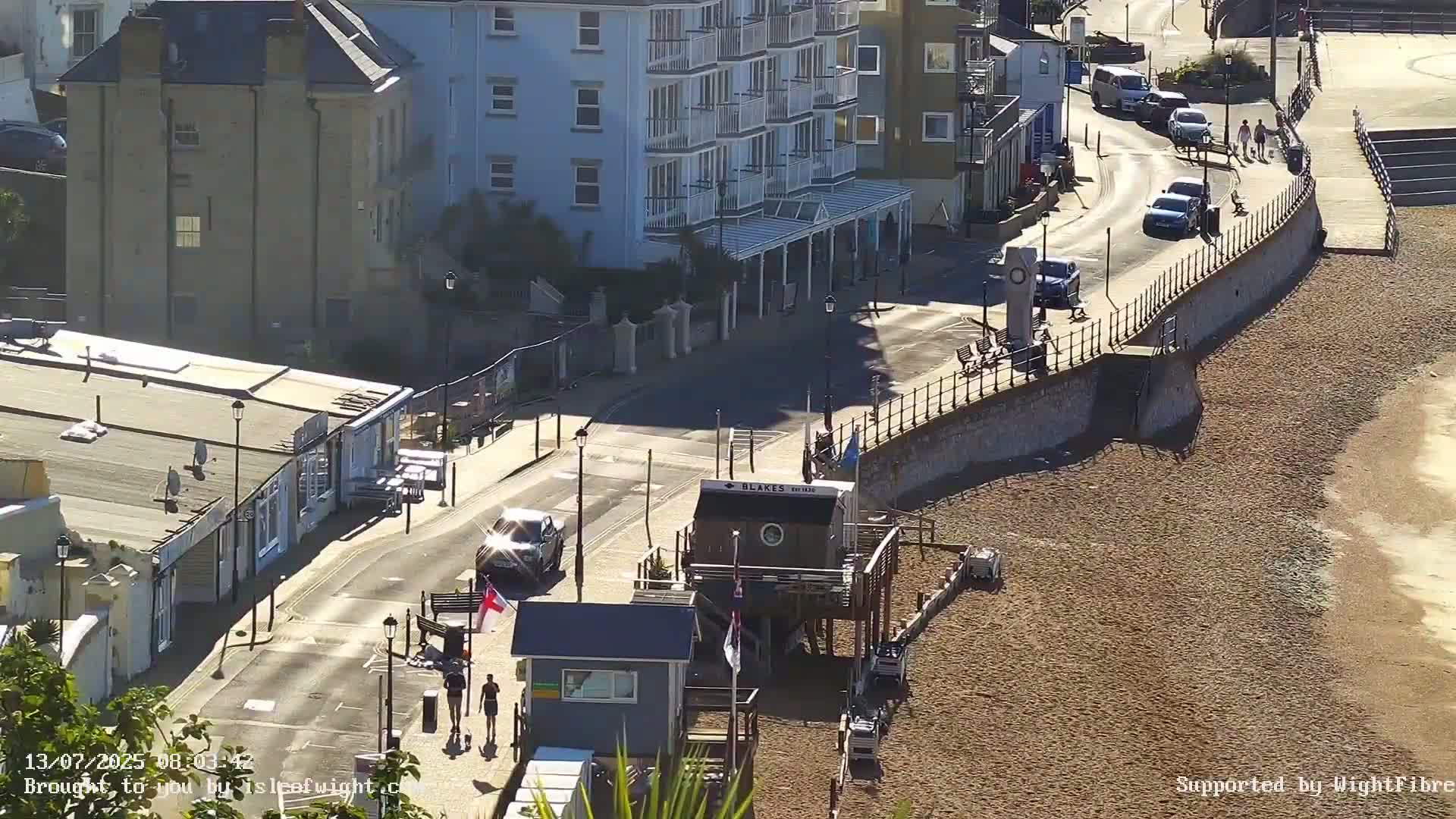 A sunny seaside town scene shows a street with parked and moving cars, buildings, a beach with a small structure on it, and pedestrians walking along a sea wall.