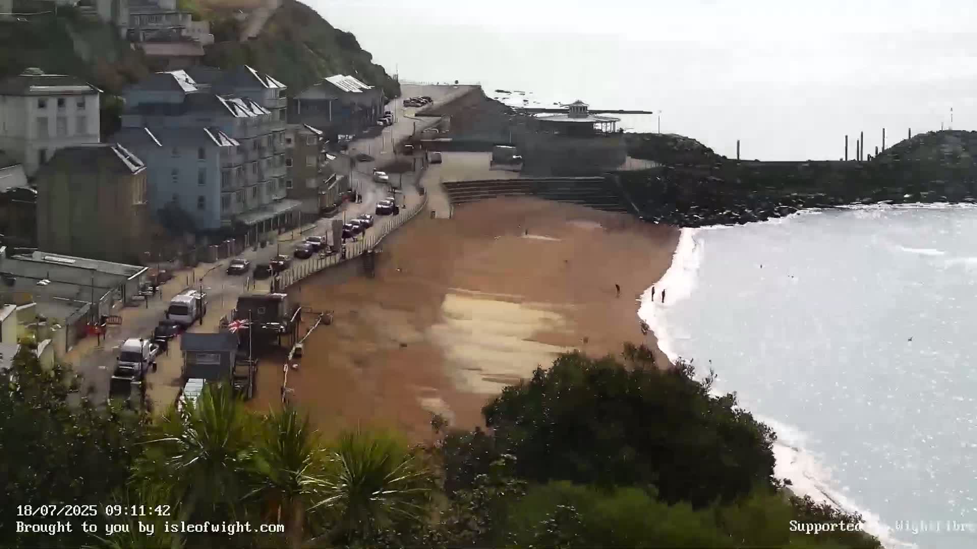A sandy beach with a few people on it is bordered by buildings on a hillside and a grayish sea with small waves under an overcast sky.
