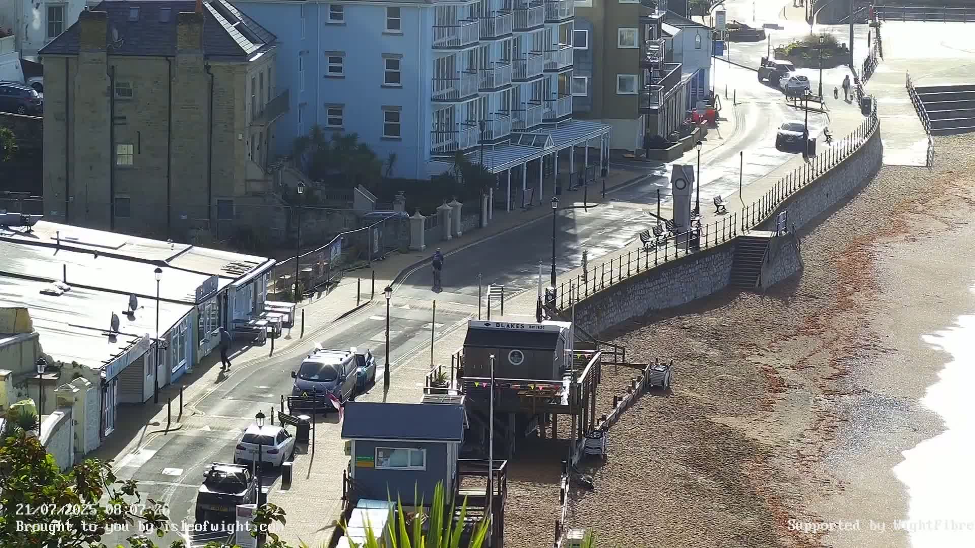 A sunny day shows a seaside town with a pebble beach, buildings, parked cars, and a few pedestrians along a road next to a seawall.