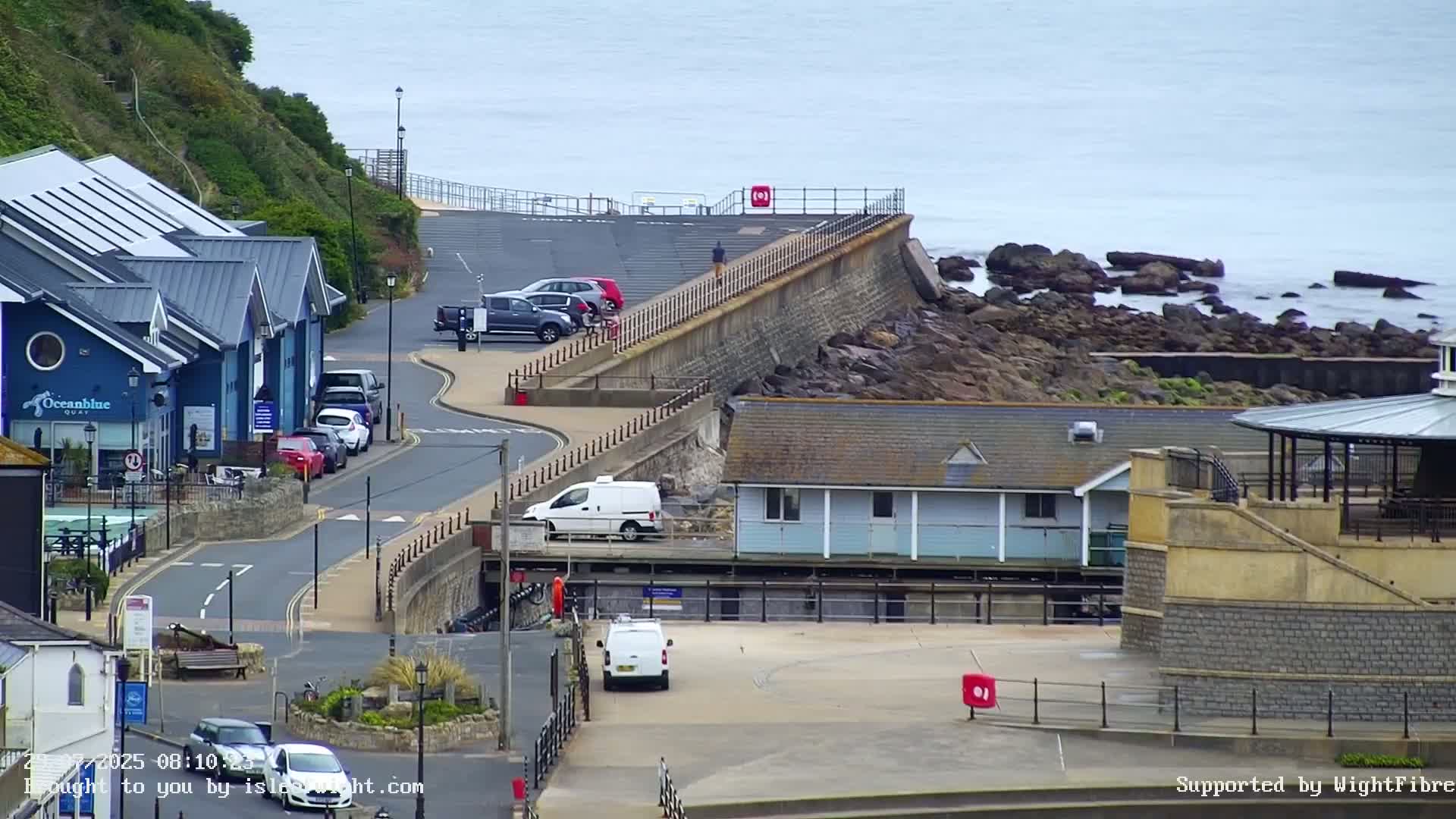 A coastal scene on an overcast day shows a road curving alongside a seawall, with buildings, parked cars, and a rocky shoreline visible.