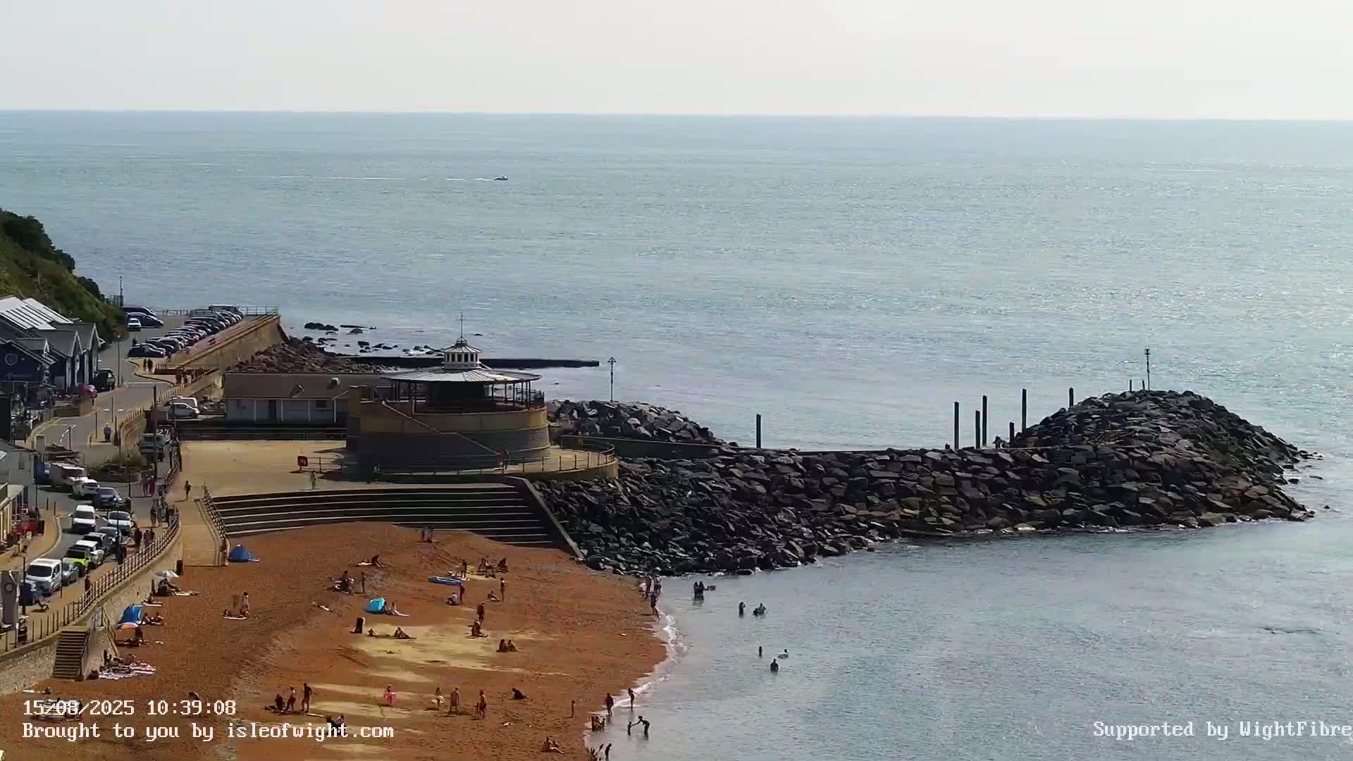 A coastal scene on an overcast day shows a road curving alongside a seawall, with buildings, parked cars, and a rocky shoreline visible.