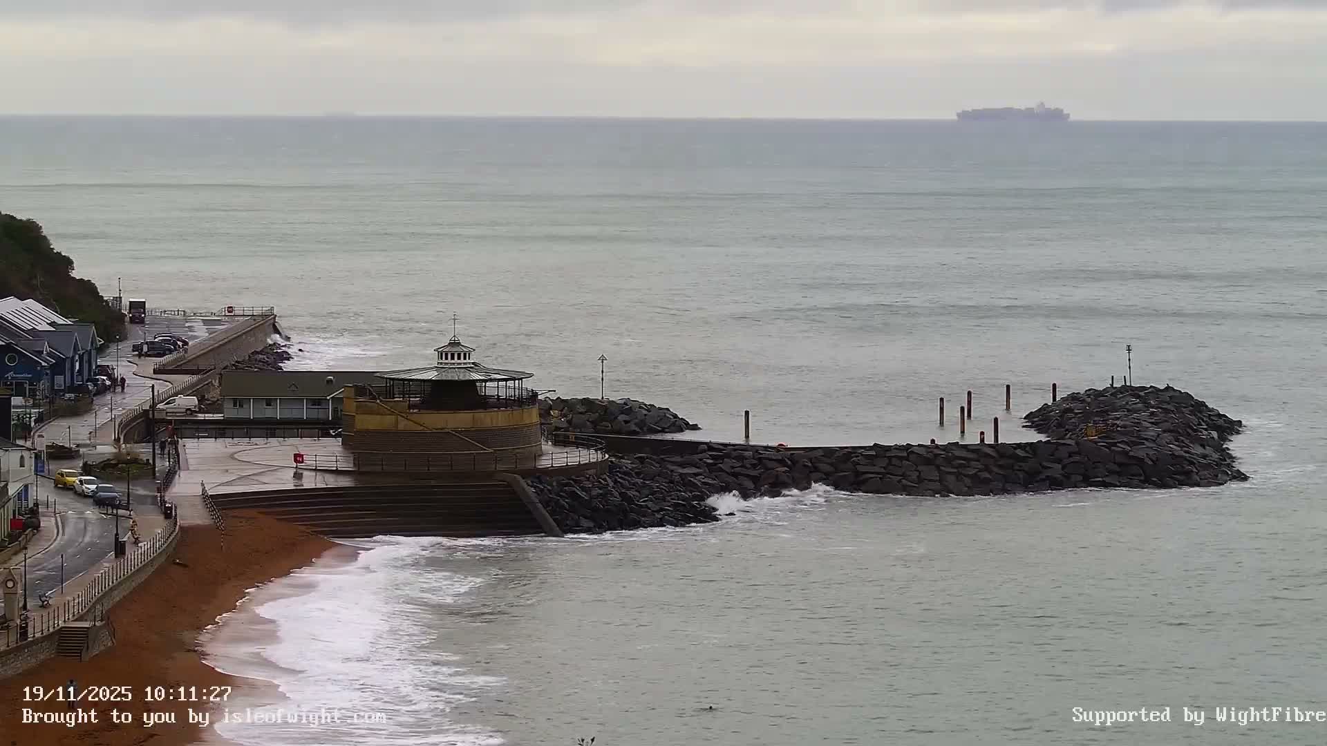 Under an overcast sky, a coastal scene features a sandy beach with breaking waves, a road winding past buildings, a prominent round building next to a series of rocky breakwaters, and a large ship on the distant, choppy sea.