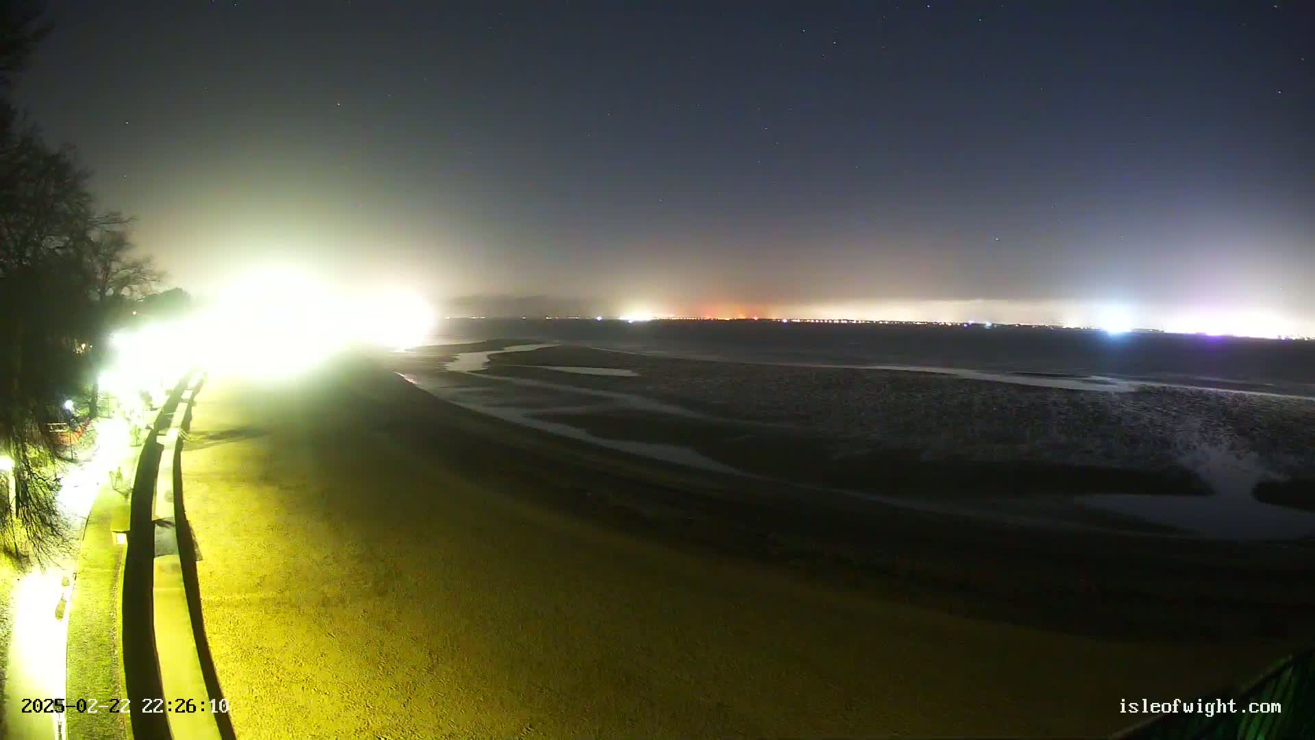 A brightly lit sandy beach at night stretches towards a dark body of water under a starry sky, with city lights visible in the distance.