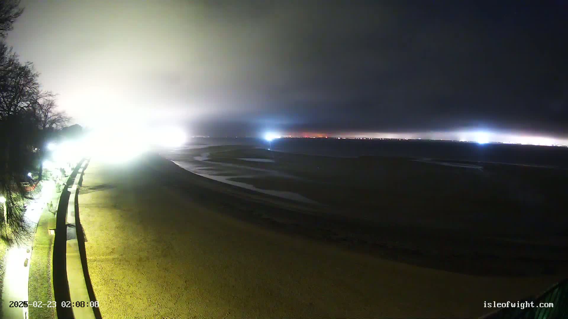 A night-time, foggy view of a beach with lights illuminating a walkway and the distant shore.