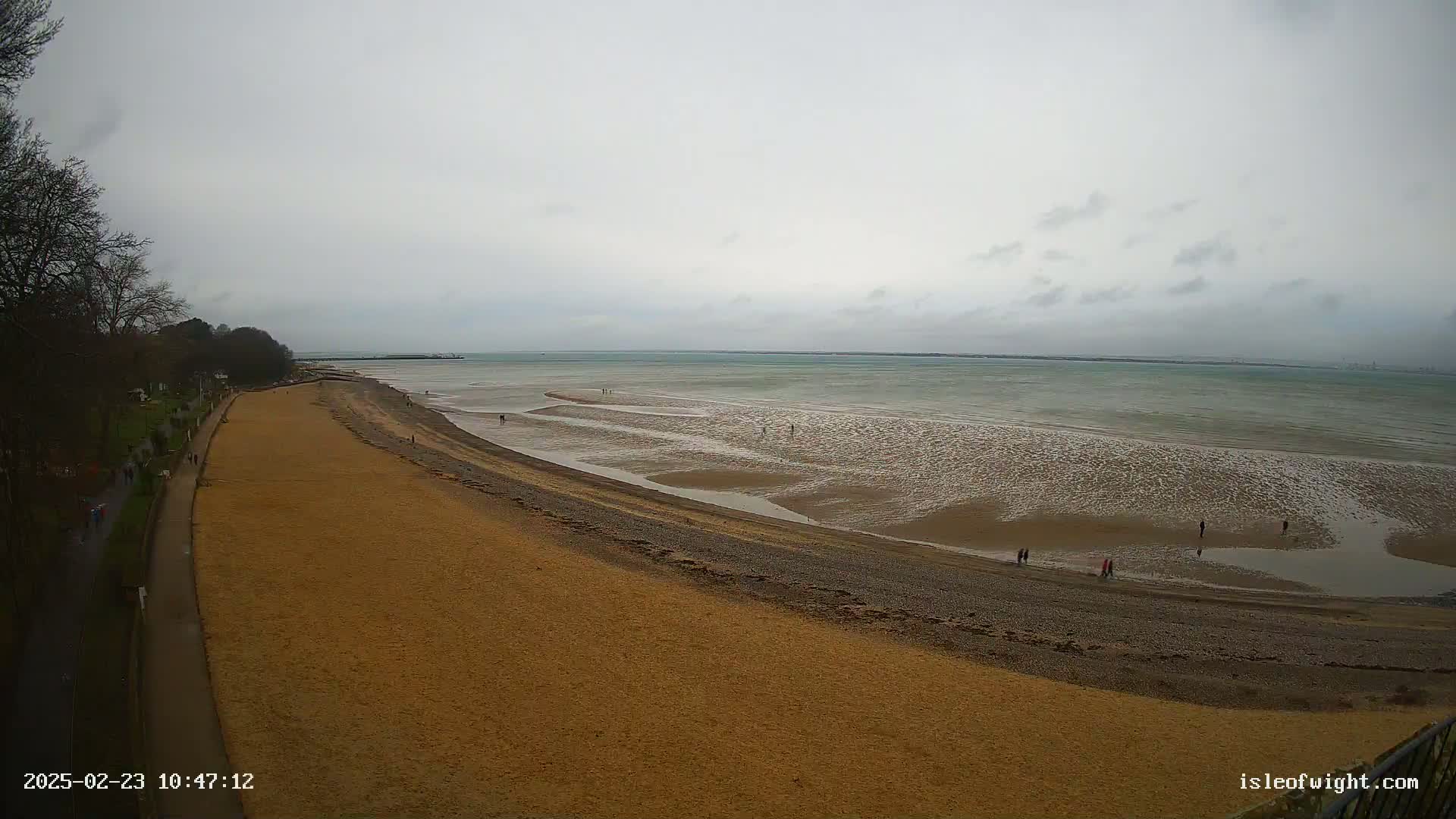 A wide sandy beach stretches along a calm, gray sea under an overcast sky, with a few people walking near the water's edge.