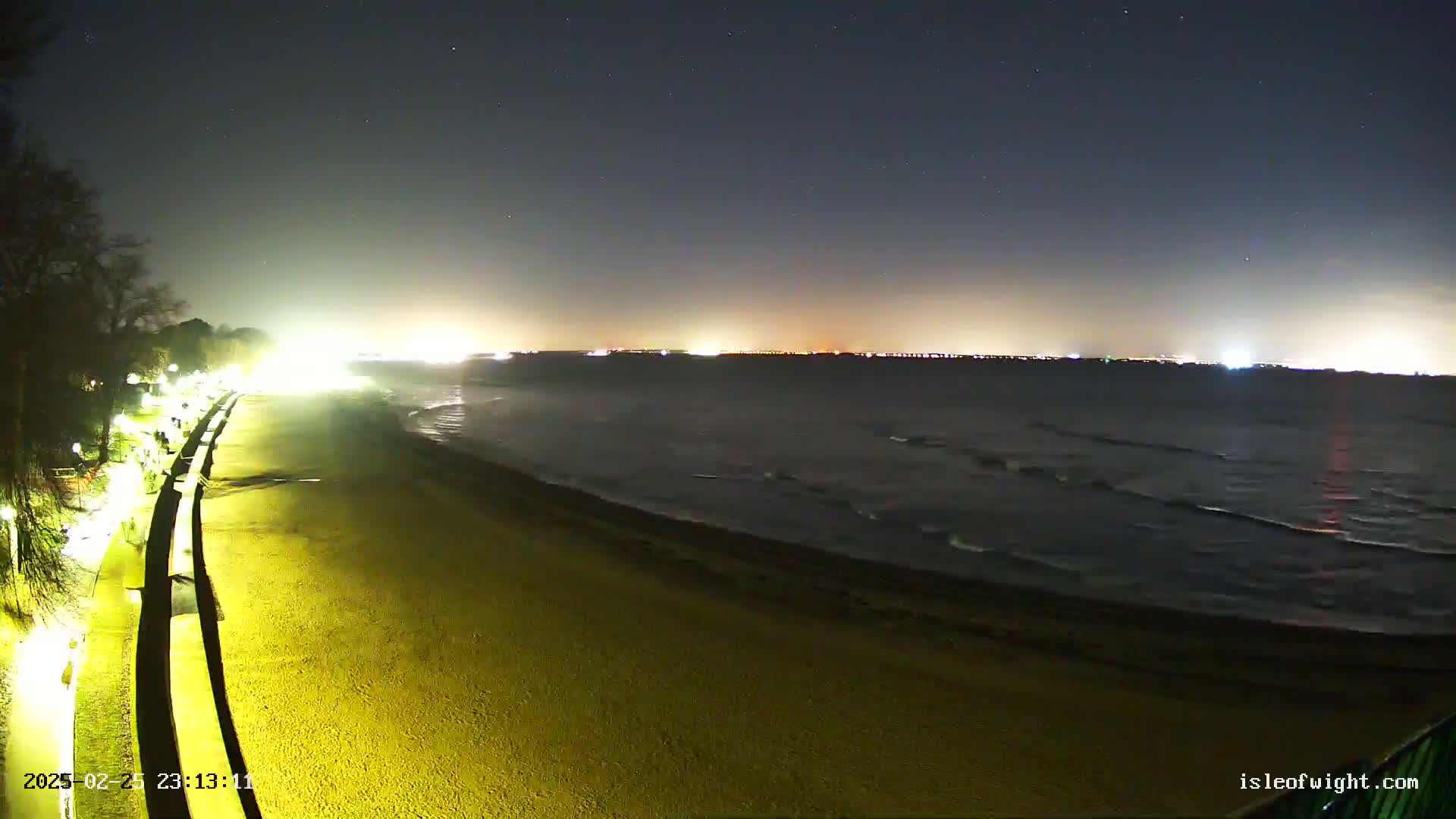 A dark, nighttime beach scene shows a gently rolling sea under a starry sky, with illuminated walkways and distant city lights visible across the water.