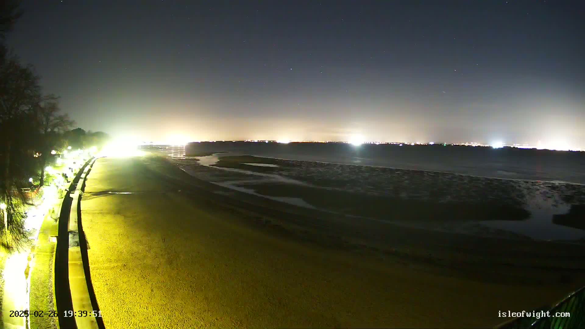 A nighttime view of a sandy beach and shoreline with lights illuminating a walkway and distant city lights under a clear, starry sky.