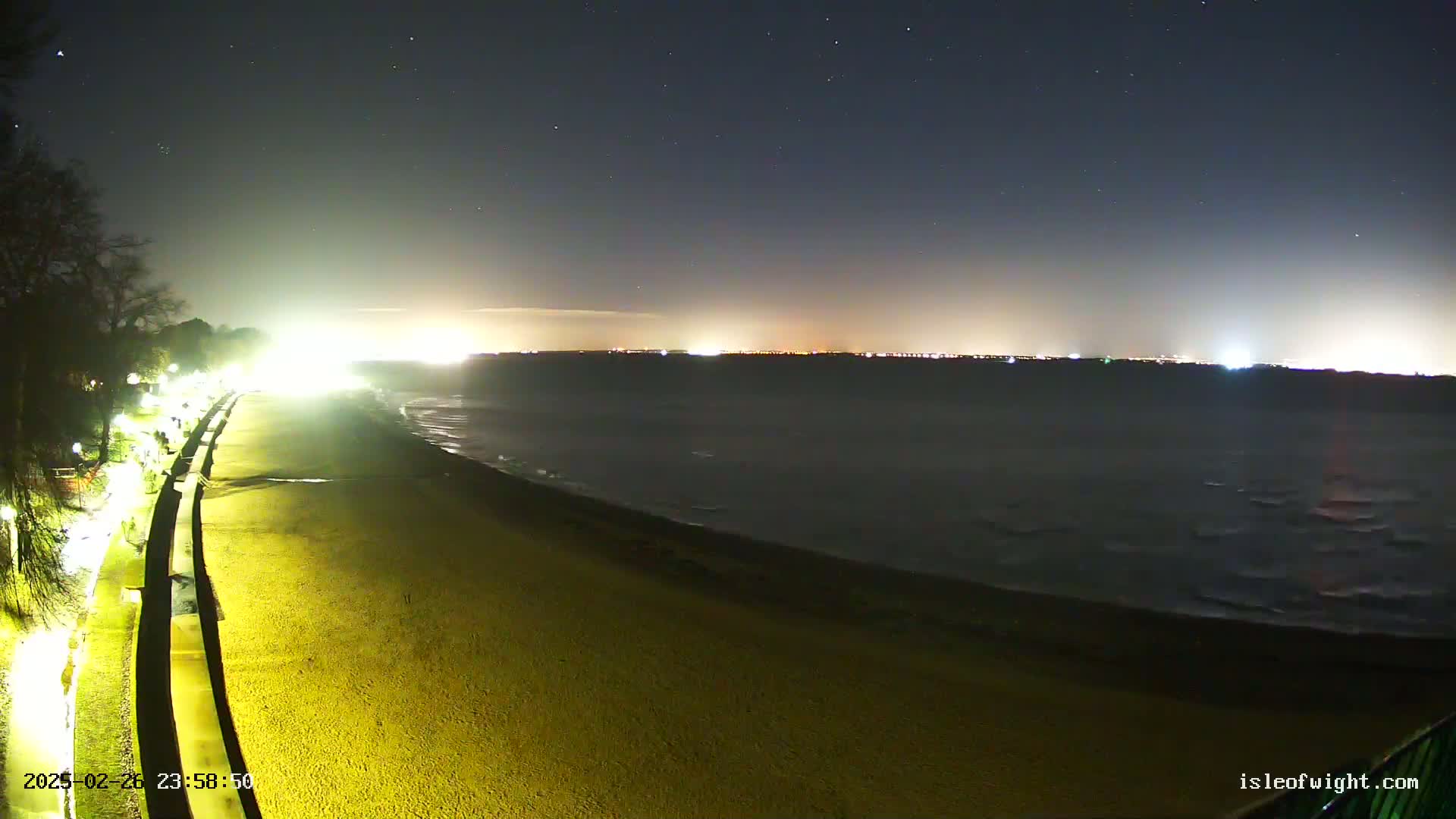 A nighttime view of a gently sloped beach next to a paved walkway, illuminated by artificial lights, with a dark, somewhat wavy body of water extending to a distant, faintly lit cityscape under a starry sky.