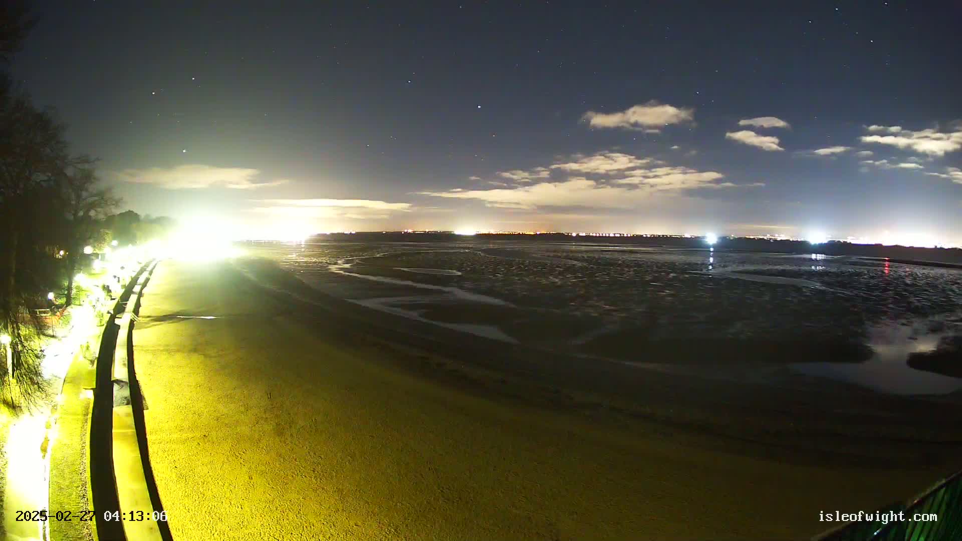A nighttime view of a beach at low tide, under a partly cloudy sky with stars visible, shows a lit walkway and distant city lights.
