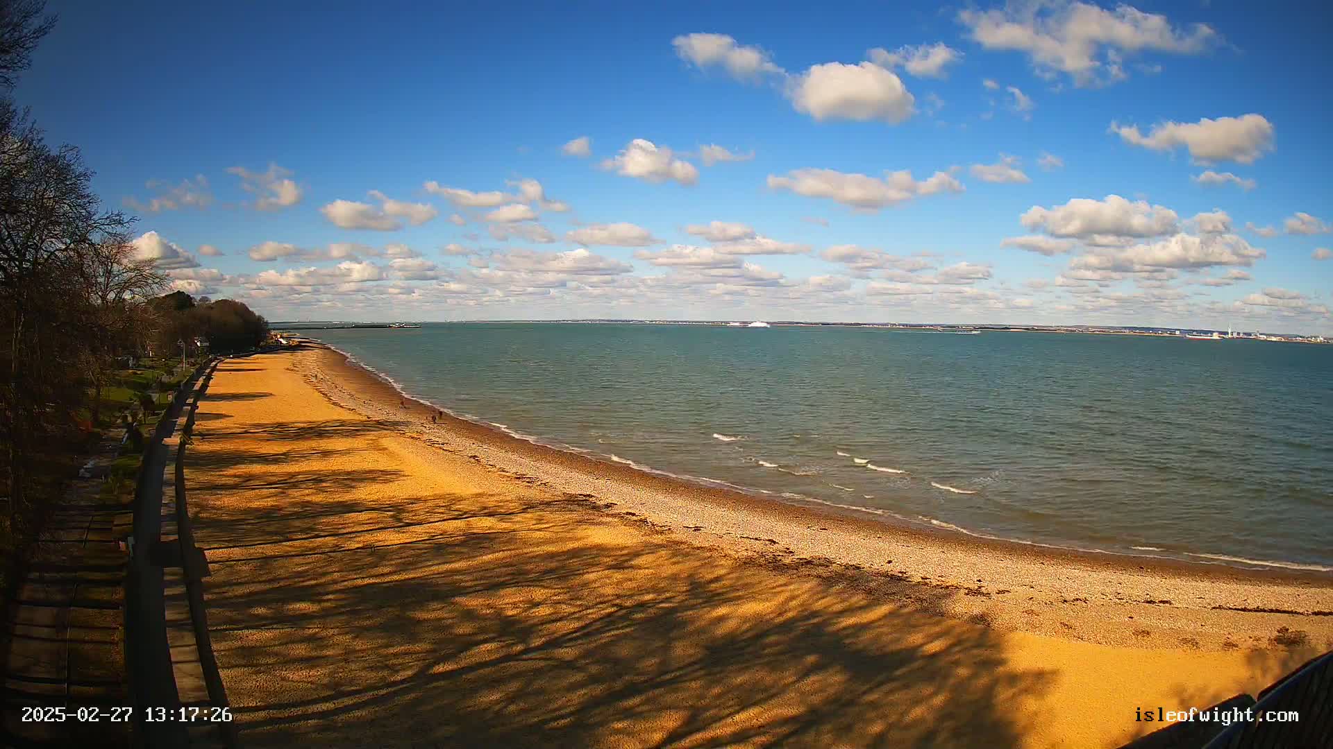 A sandy beach stretches along a calm sea under a mostly sunny sky with scattered cumulus clouds.