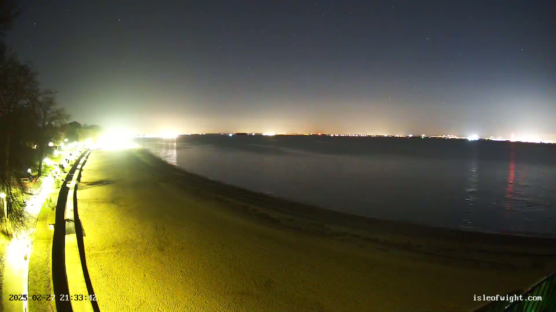 A nighttime view of a beach with a dimly lit shoreline and a body of water reflecting distant city lights under a clear, starry sky.