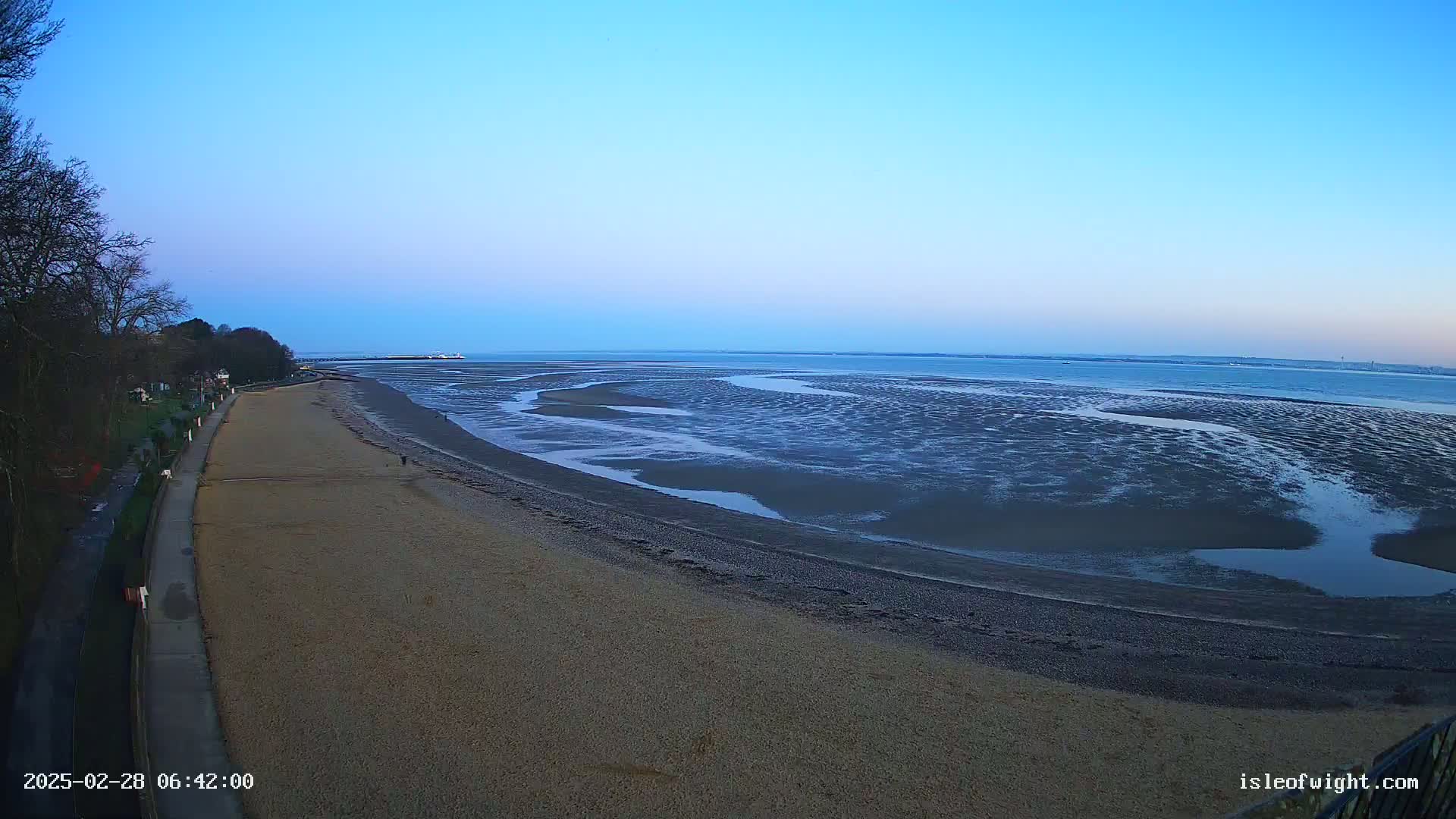 A sandy beach stretches alongside a calm, blue body of water at low tide under a clear, twilight sky.