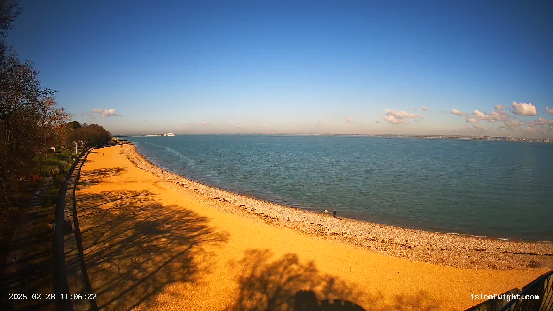 A sandy beach curves along a calm, blue sea under a mostly clear, sunny sky.