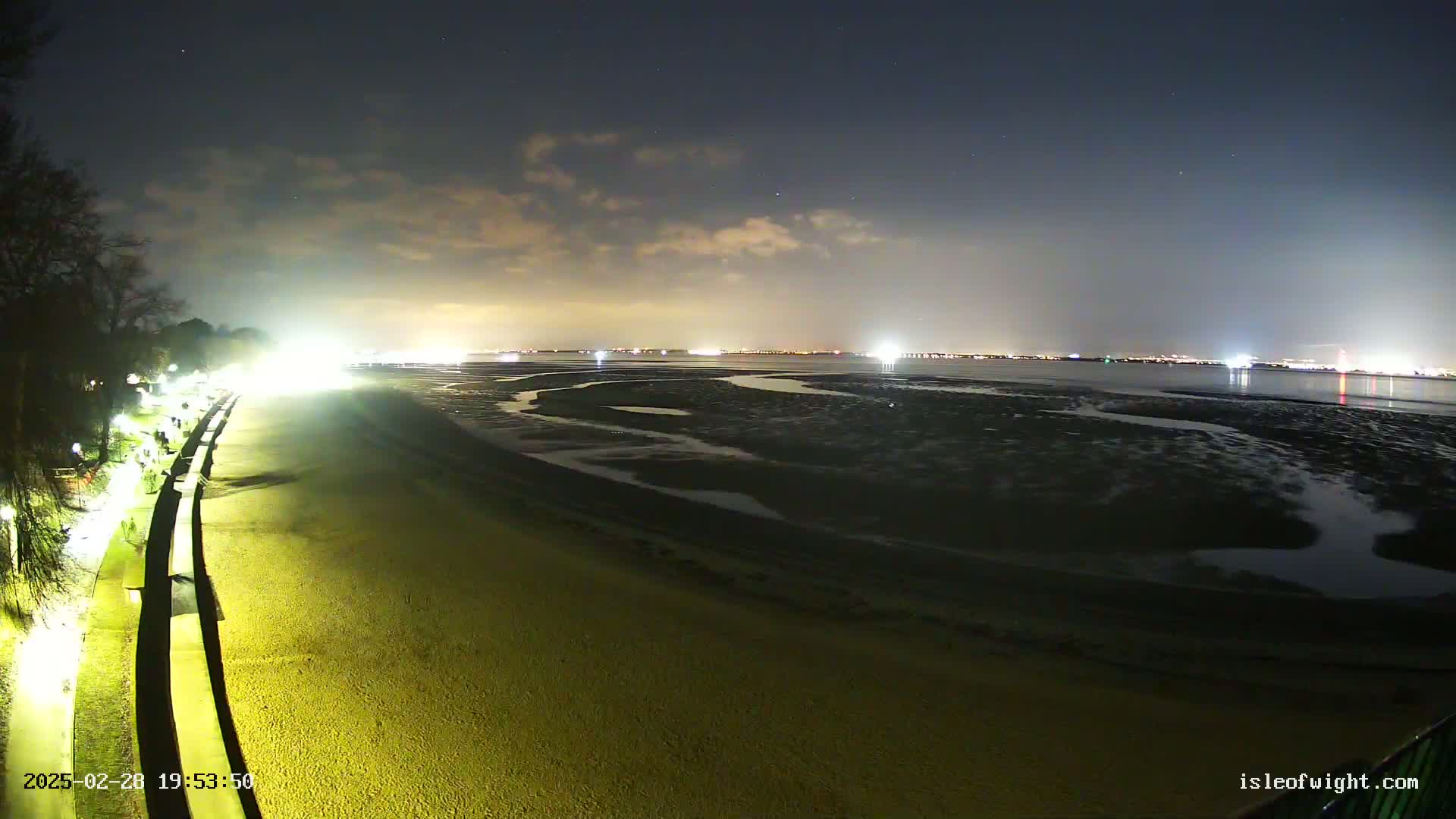 A nighttime view of a sandy beach illuminated by artificial lights, with a calm body of water revealing tidal flats and a distant, faintly lit cityscape under a partly cloudy sky.