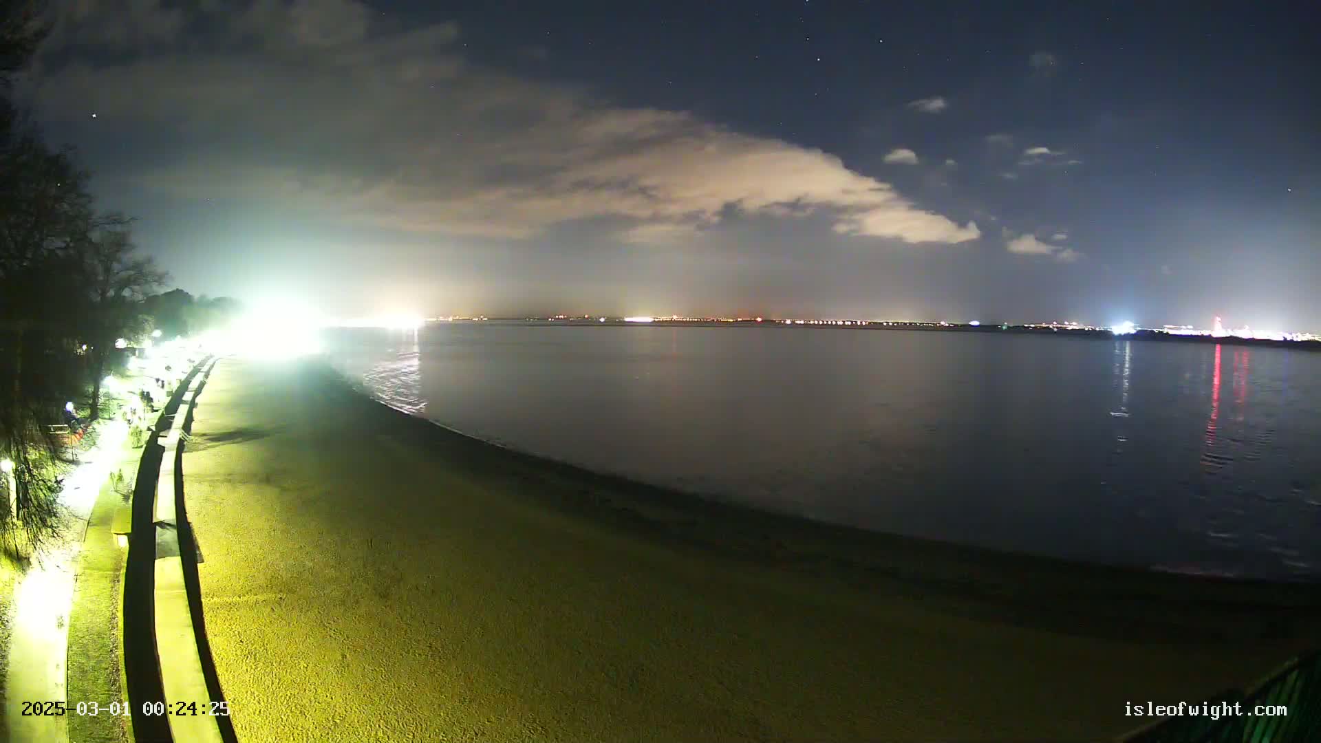 A nighttime view of a sandy beach with a lit promenade, gently lapping waves, and a distant city skyline under a partly cloudy sky.