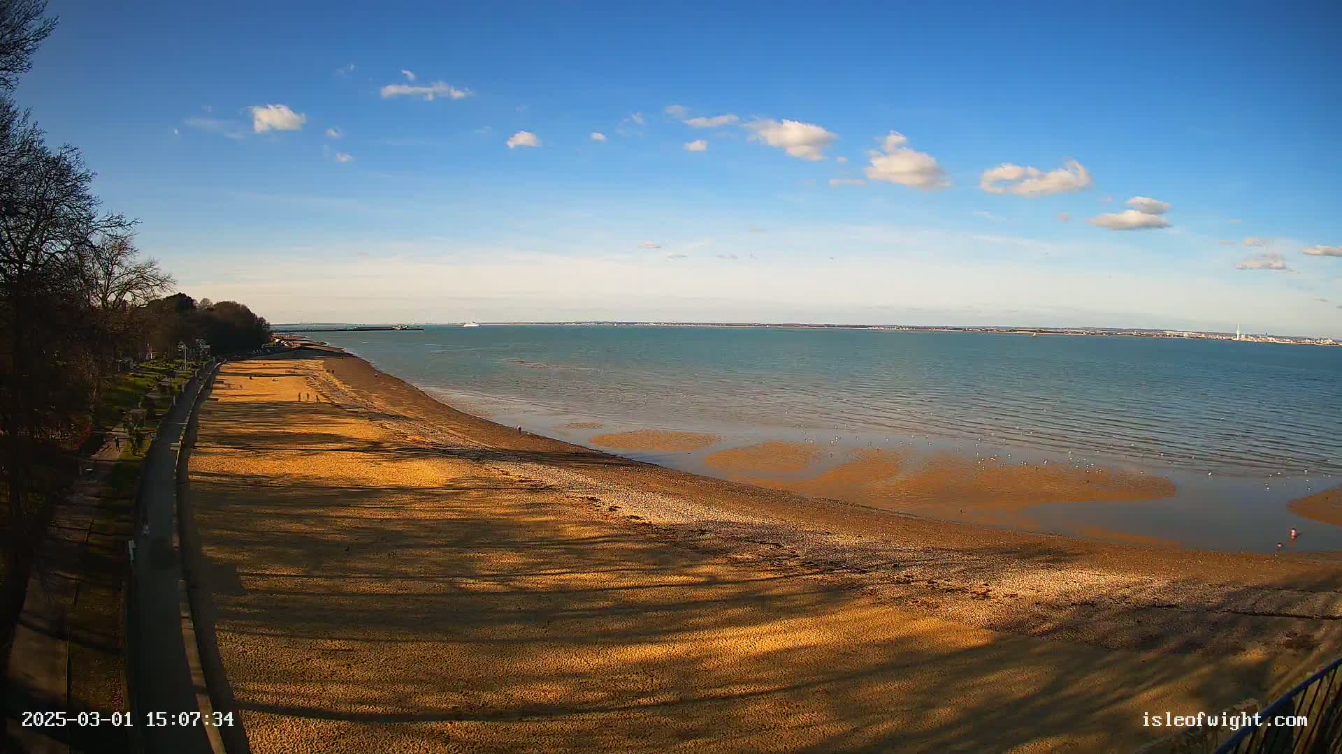 A sandy beach stretches along a calm body of water under a mostly clear blue sky with a few scattered clouds.