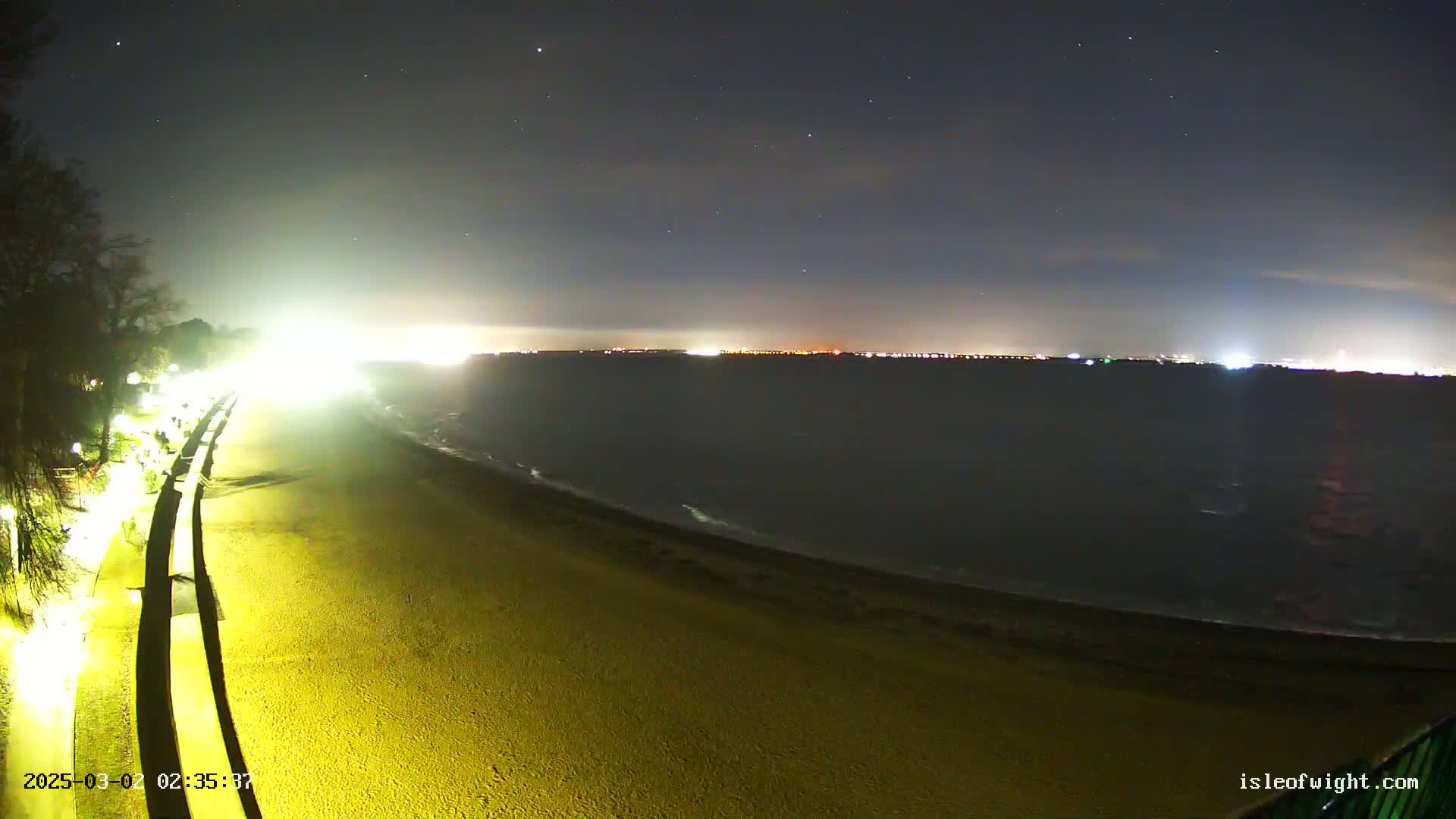 A nighttime view of a sandy beach with a paved walkway lined with lights, overlooking a dark body of water with a distant, faintly lit cityscape under a clear, starry sky.