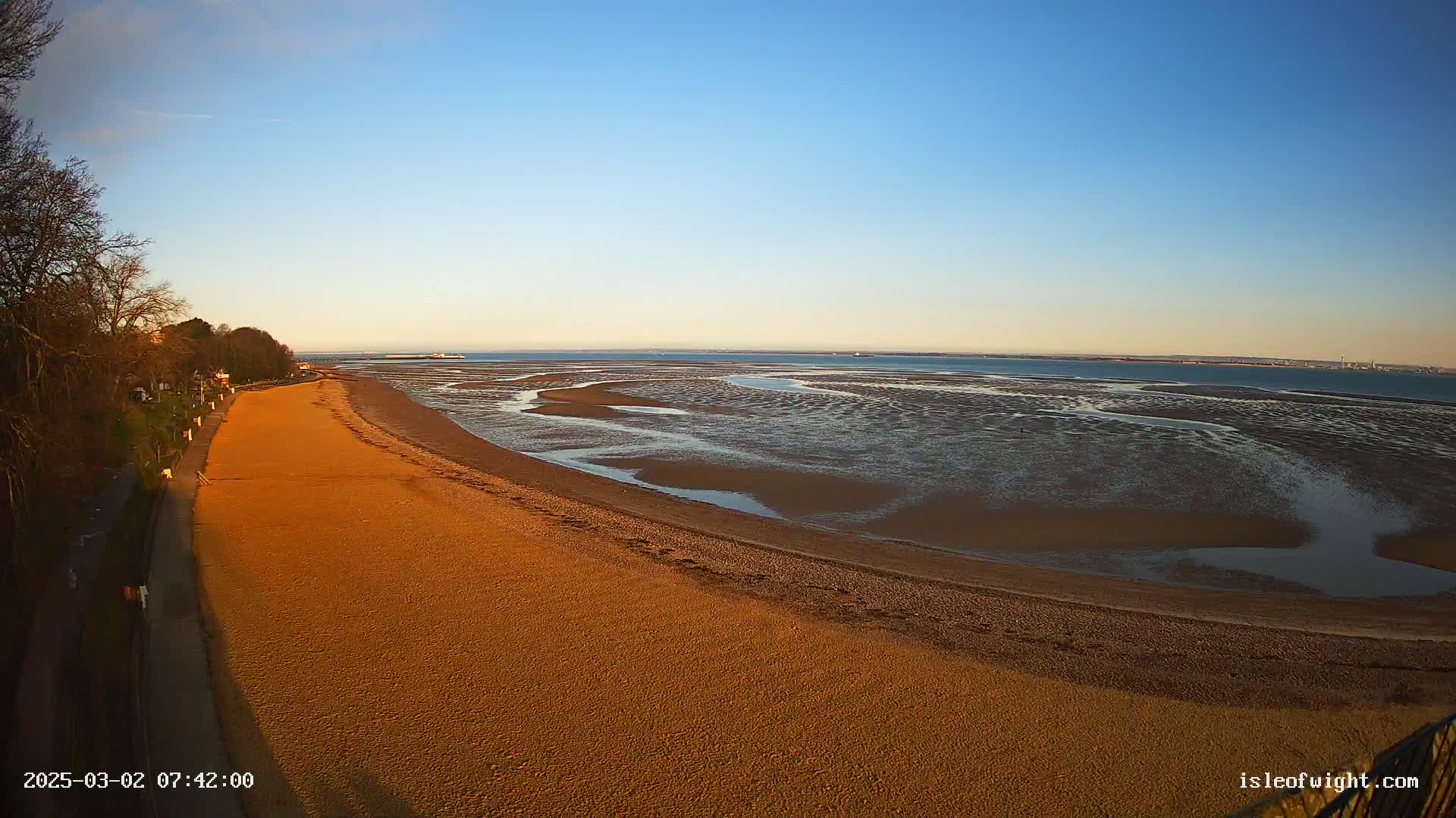 A long sandy beach stretches out to a calm, wide expanse of exposed mudflats and water under a clear blue sky.
