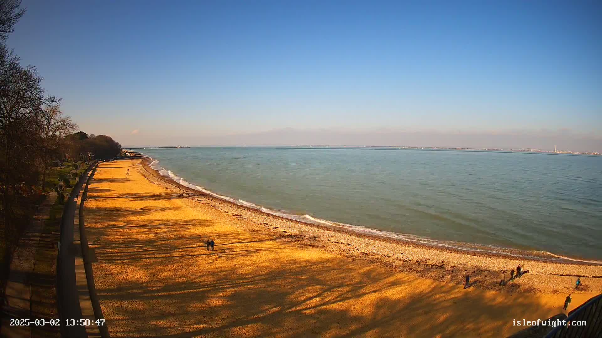 A sandy beach curves along a calm, blue-green sea under a clear blue sky, with a few people scattered along the shore and tree shadows cast on the sand.