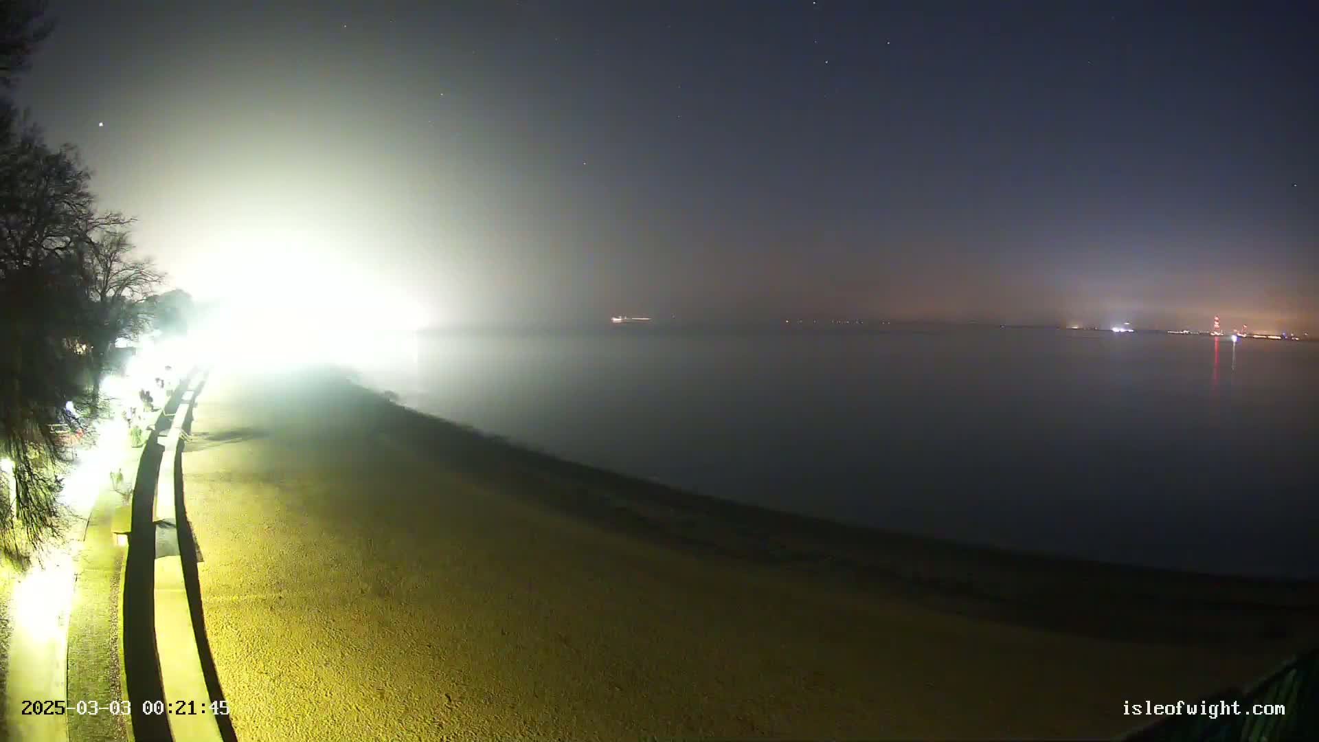 A fog-shrouded beach at night is illuminated by artificial lights, with a calm body of water extending to a distant shoreline.