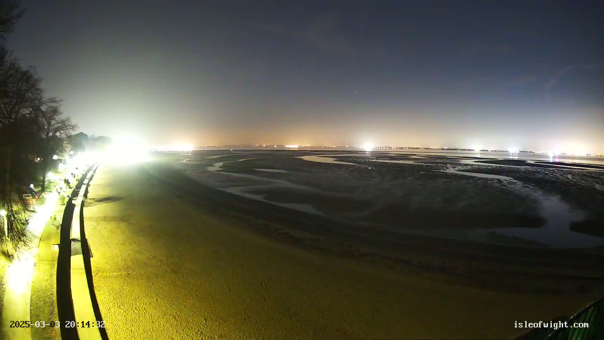 A brightly lit beach at night stretches toward a dark, starry sky over a tidal flat and distant city lights.