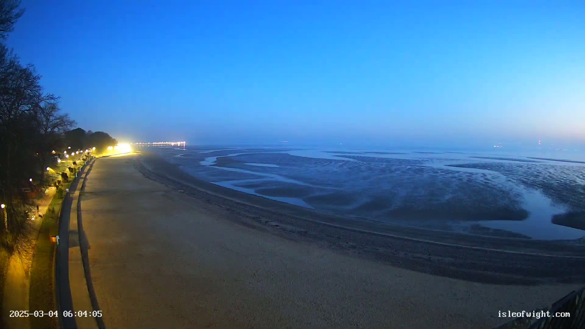 A long, sandy beach stretches out to a calm, dark blue sea at twilight, illuminated by lights along a seaside promenade.