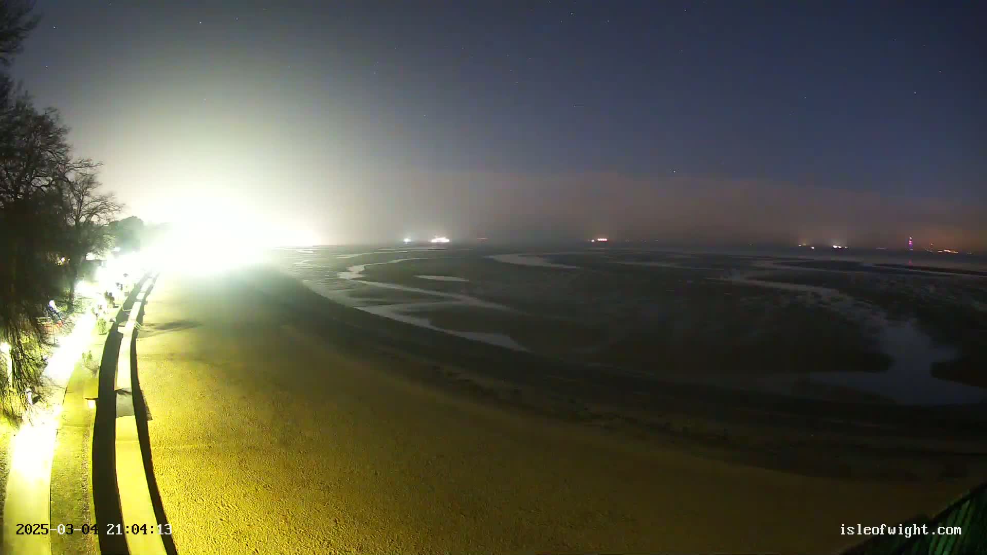 A brightly lit sandy beach at night extends to a hazy, dark body of water under a starry sky.