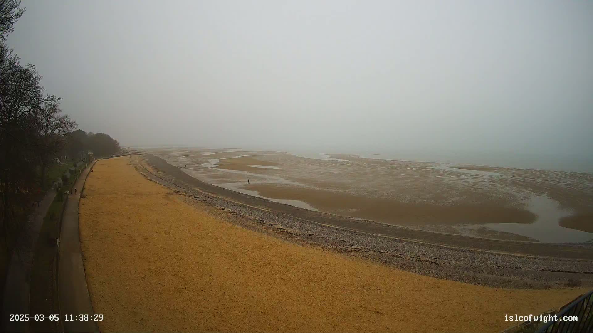 A sandy beach curves alongside a calm, foggy sea, with a few people walking along the shore and a paved walkway visible beside the beach.