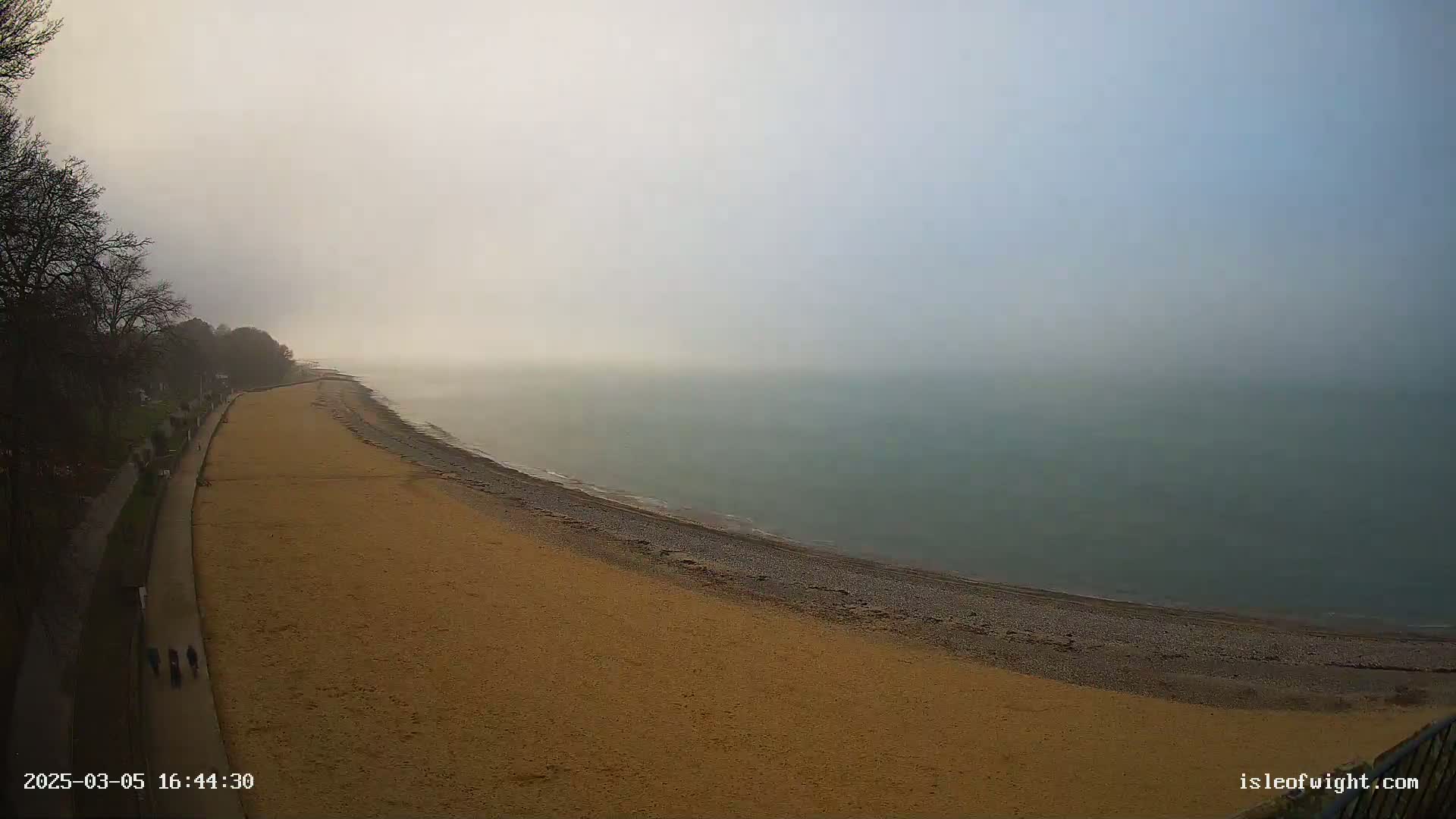 A foggy view of a sandy beach curving along a calm sea, with a paved walkway and trees visible in the background.