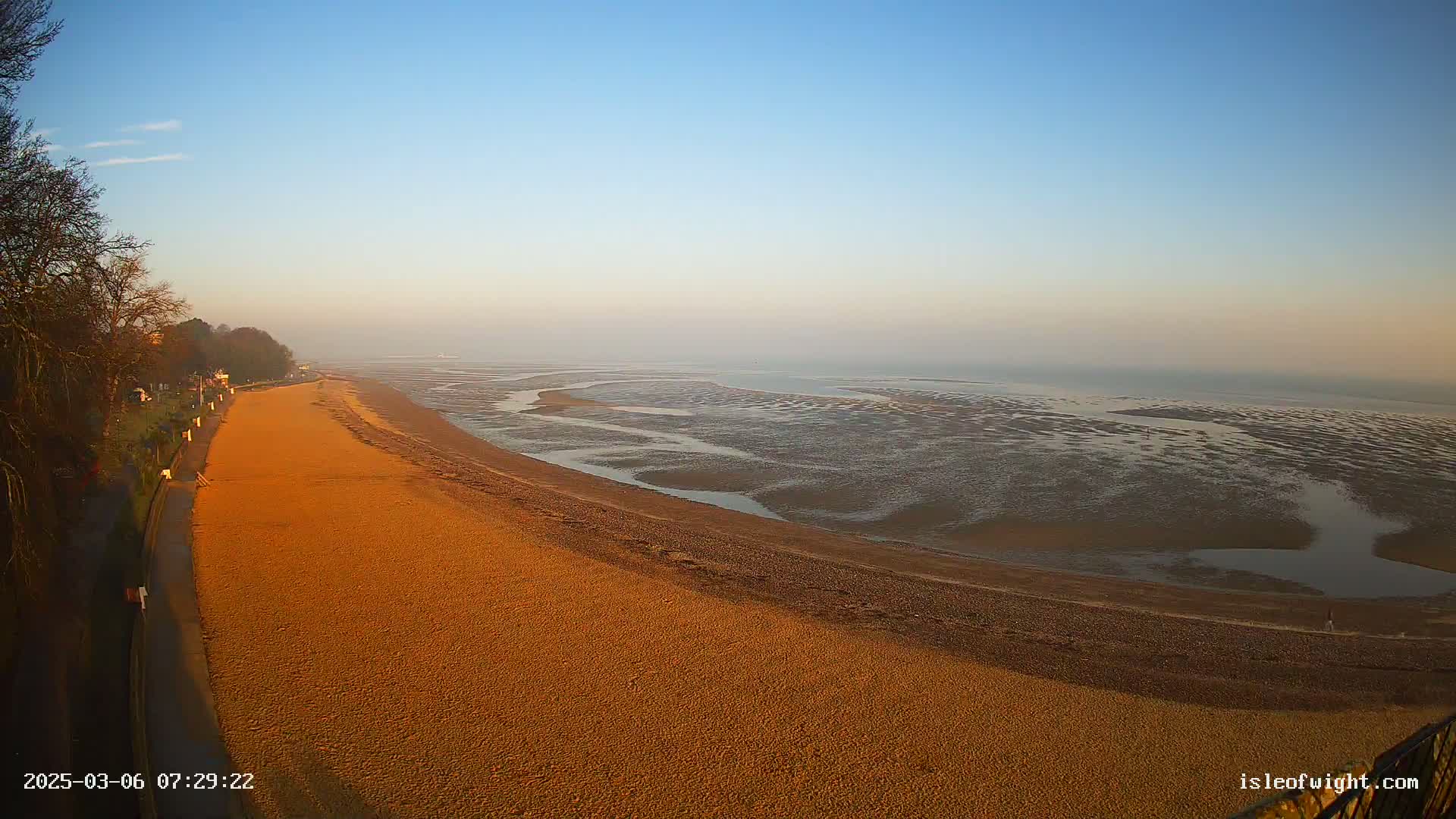 A long, sandy beach stretches along a coastline under a clear, light blue sky at dawn, with a low-lying fog in the distance over the exposed mudflats of the receding tide.