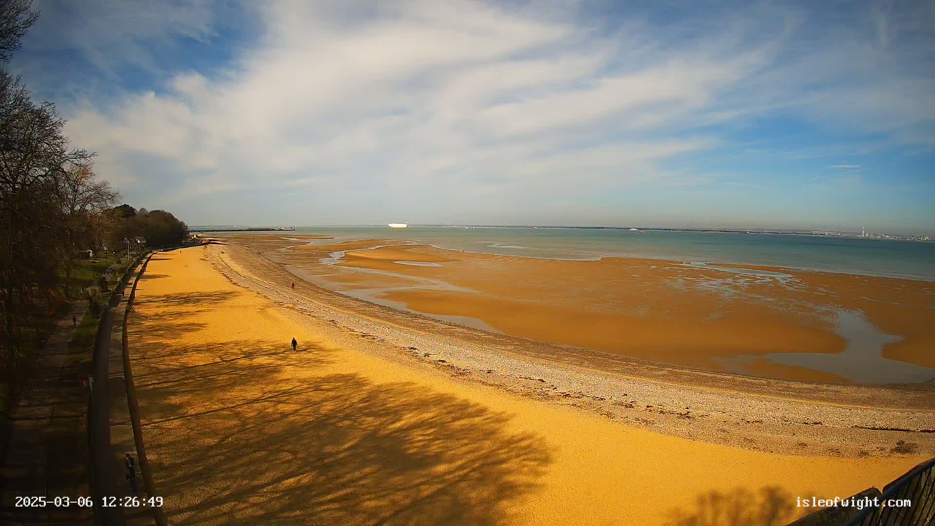 A sandy beach stretches along a calm, light-blue ocean under a partly cloudy sky, with trees and a walkway visible on the left.