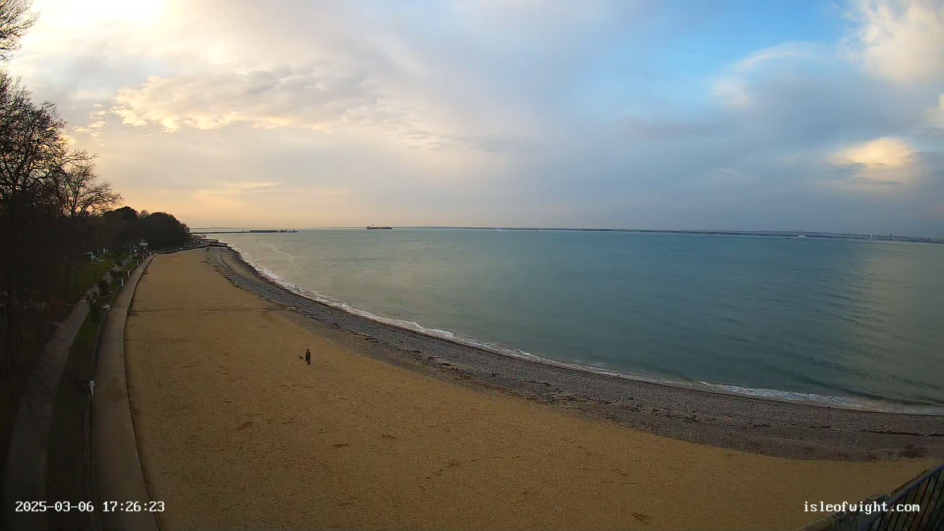 A sandy beach curves gently into a calm sea under a partly cloudy sky, with a single person walking on the beach.