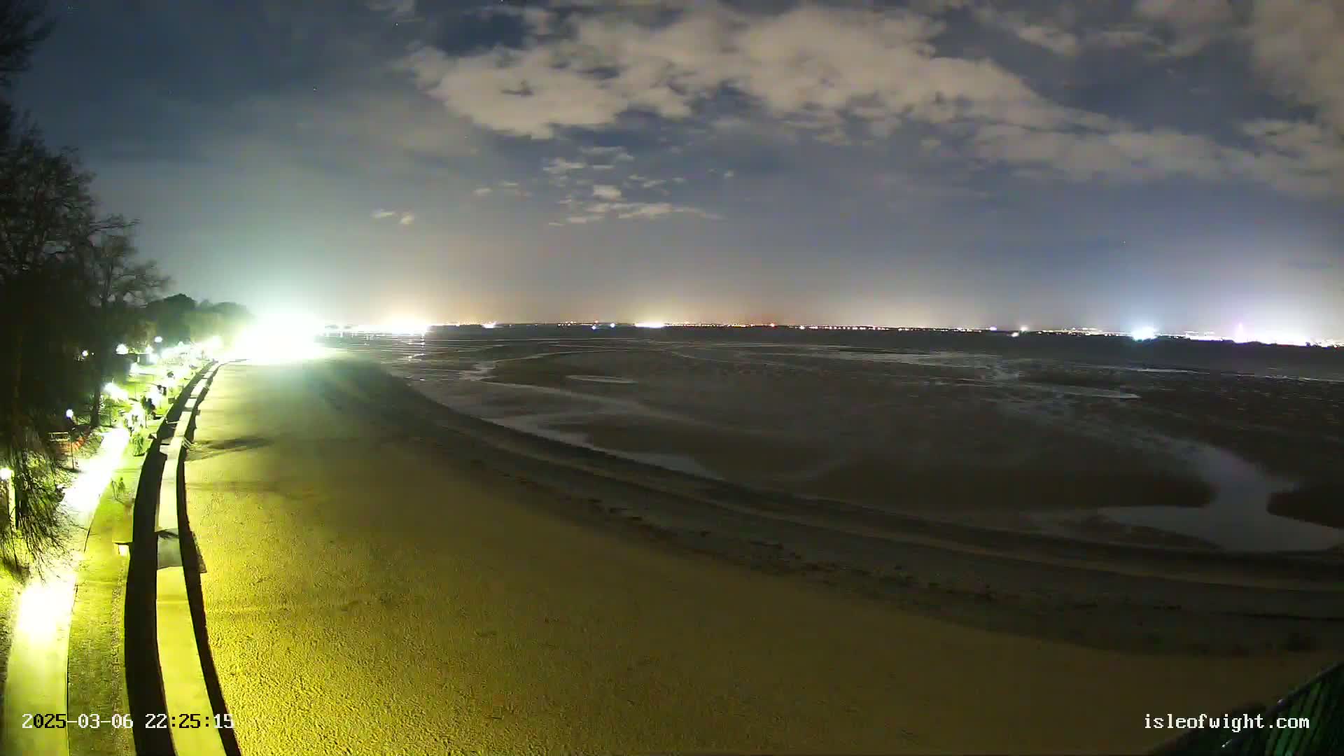 A nighttime view of a mostly empty beach, illuminated by artificial lights, with a dark, cloudy sky and distant city lights visible across the water.