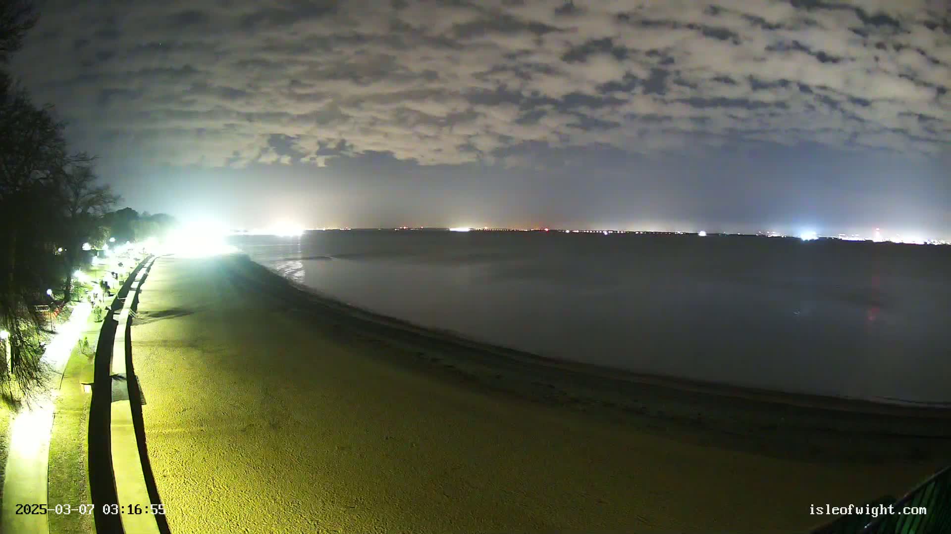 A nighttime view of a beach with a paved walkway and lights, overlooking a calm body of water with a distant city skyline under a cloudy sky.