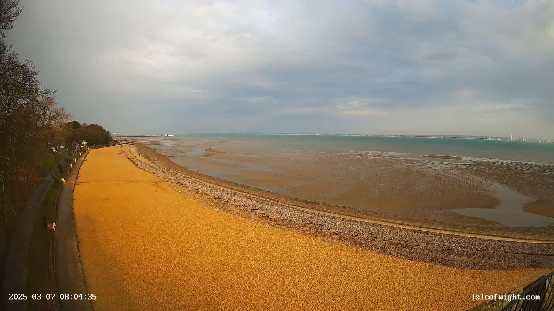 A mostly sunny day reveals a long sandy beach curving toward a calm, wide expanse of water under a partly cloudy sky.