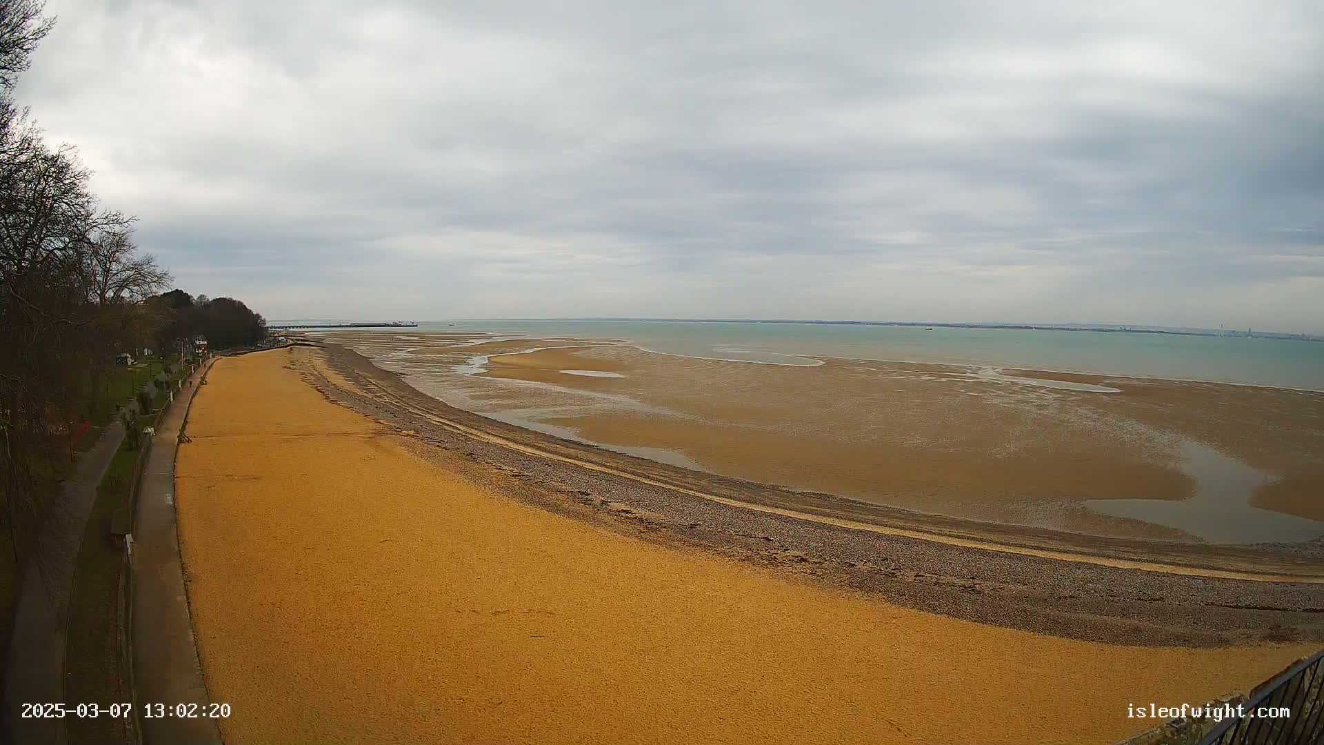 A sandy beach curves along a calm, gray-skied sea under overcast conditions, with a paved walkway visible next to the beach and some trees in the background.