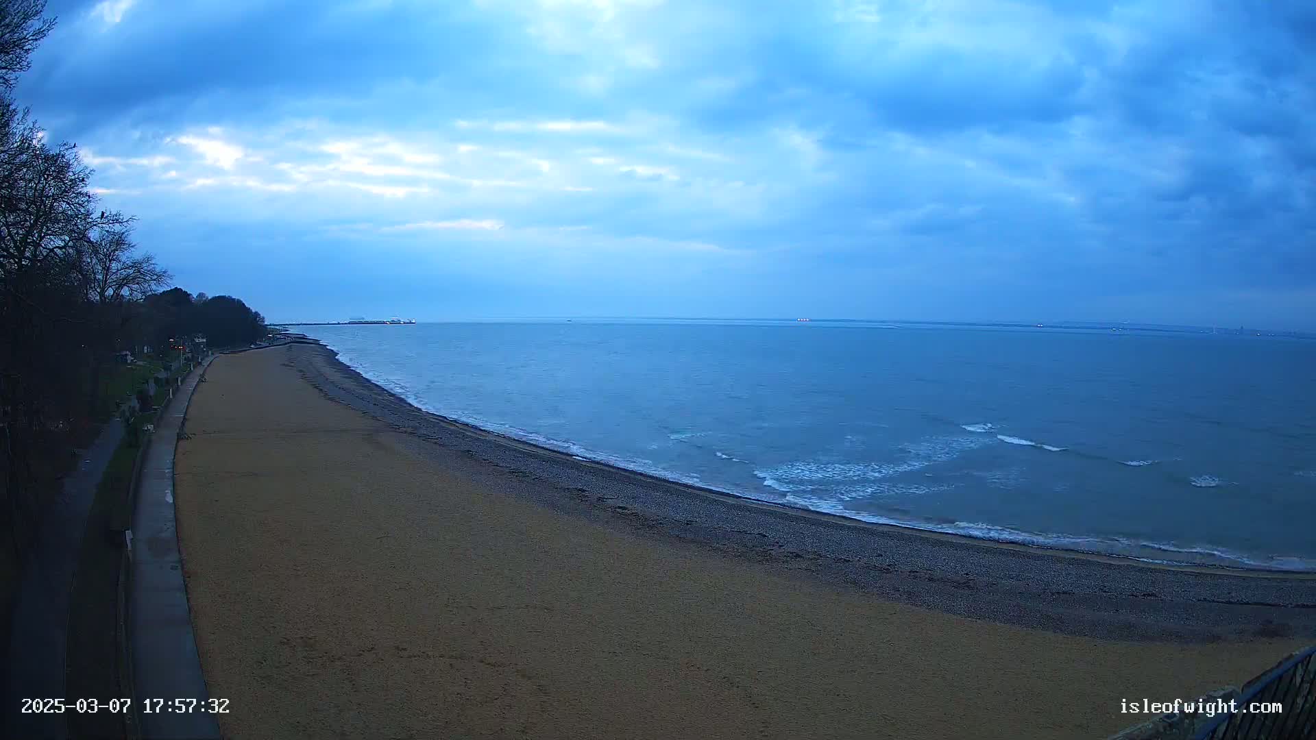 A sandy beach curves along a calm, blue sea under a mostly cloudy, dusky sky.