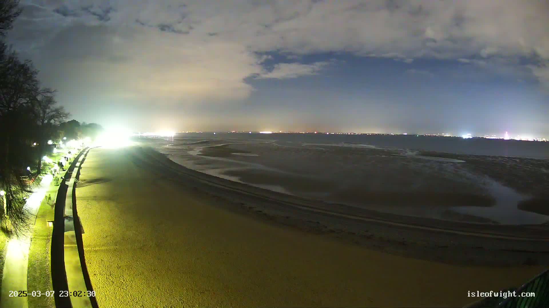 A nighttime view of a sandy beach with a paved walkway lined with lights, extending towards a calm, dark body of water under a cloudy sky.