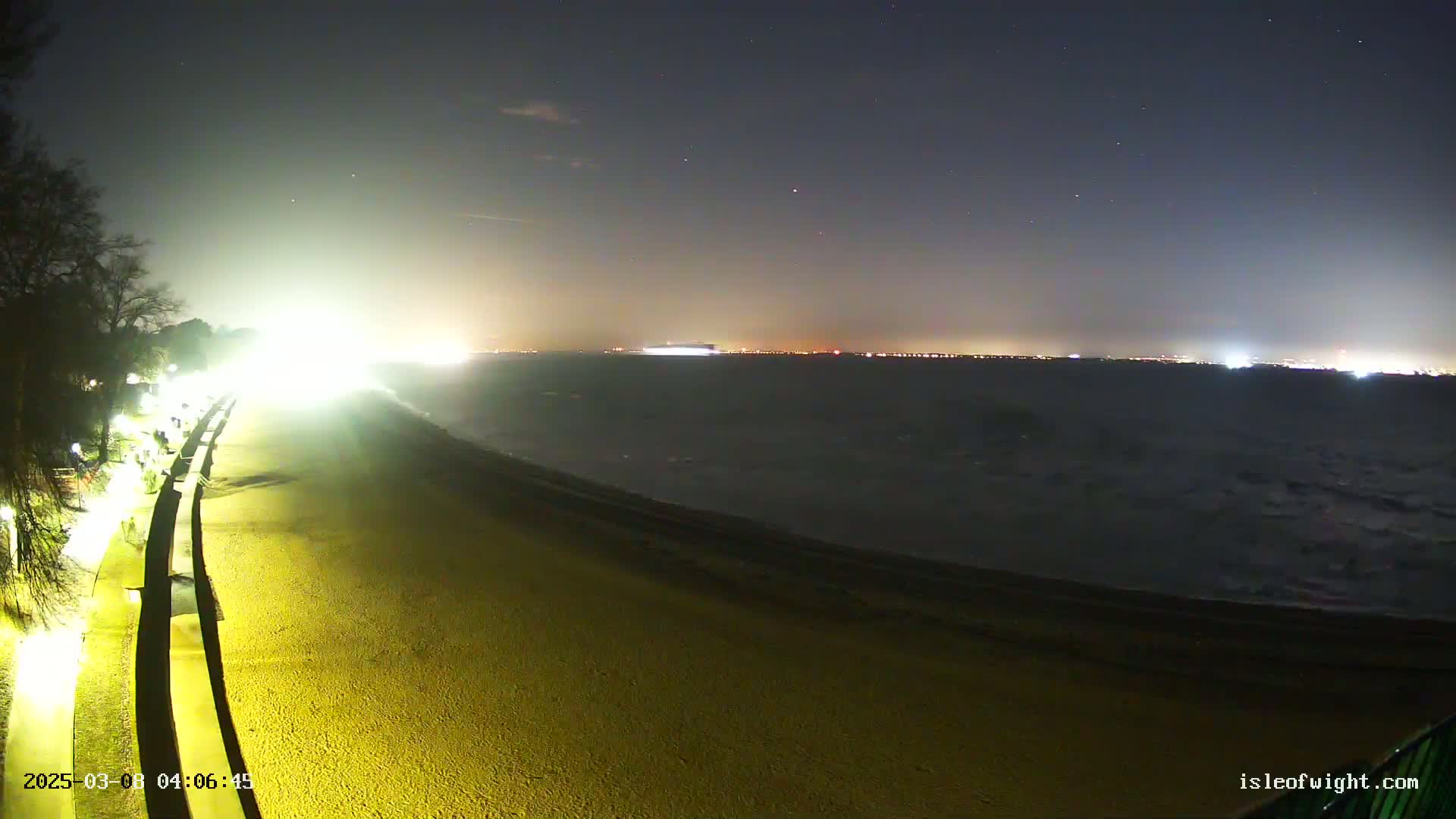 A brightly lit beach at night shows a dark, wavy ocean under a starry sky with distant city lights.