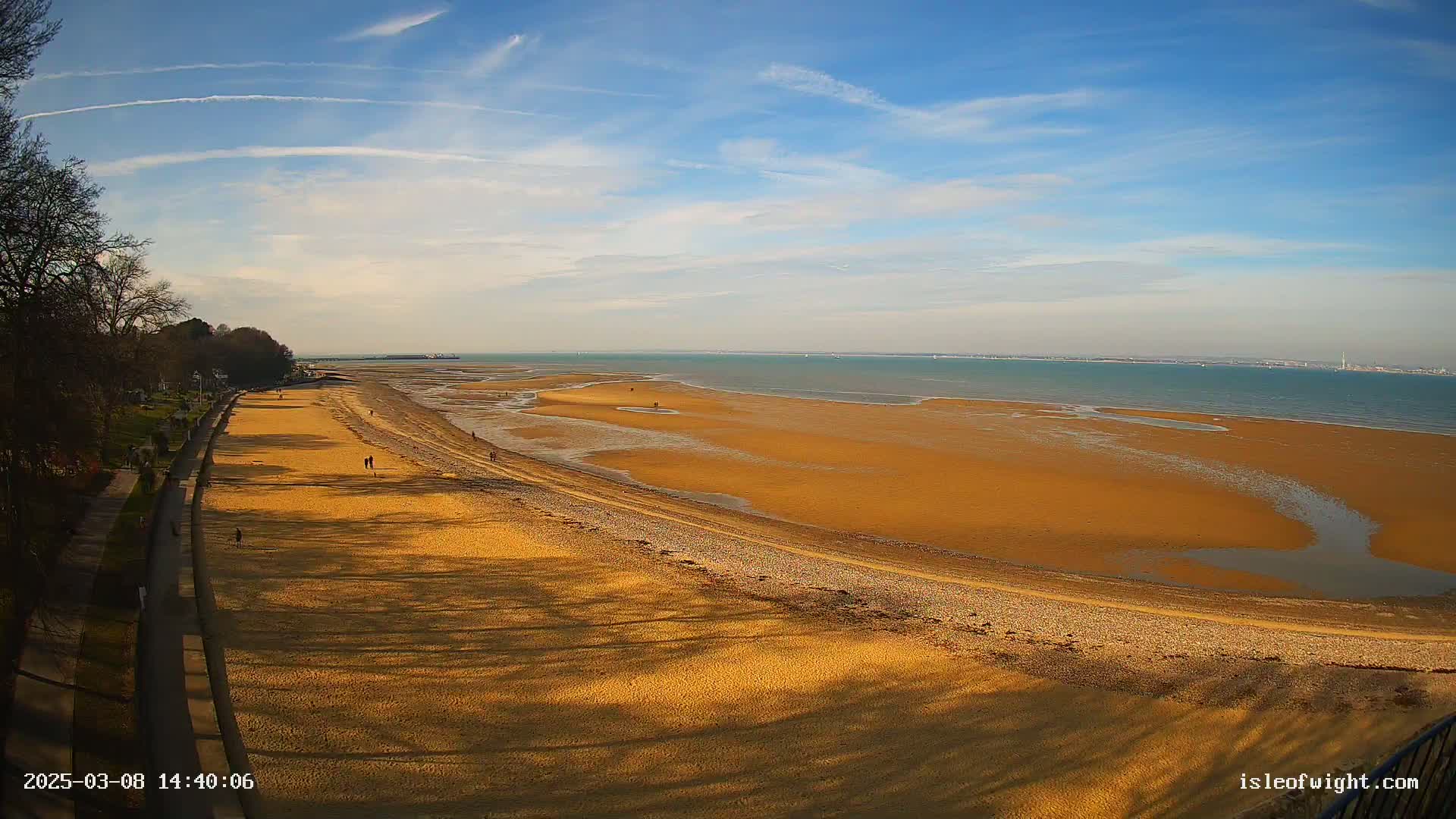 A long, sandy beach stretches out to a calm, blue sea under a mostly clear, sunny sky.
