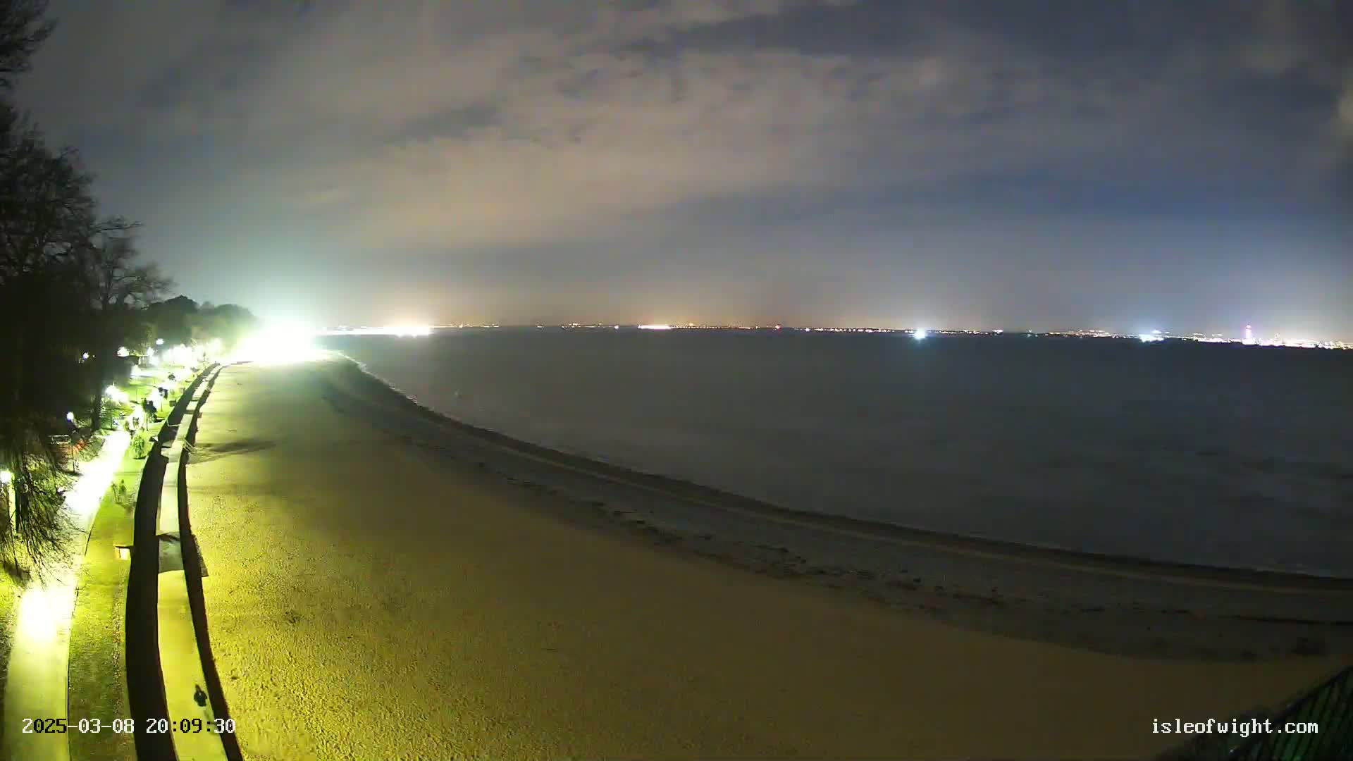 A nighttime view of a mostly empty sandy beach, a paved walkway lined with lights, and a dark body of water with a distant city skyline visible under a cloudy sky.