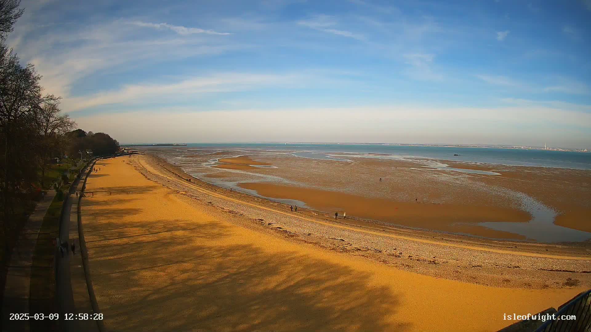 A long, sandy beach stretches out to a calm, blue sea under a mostly sunny sky, with people walking along the shore.