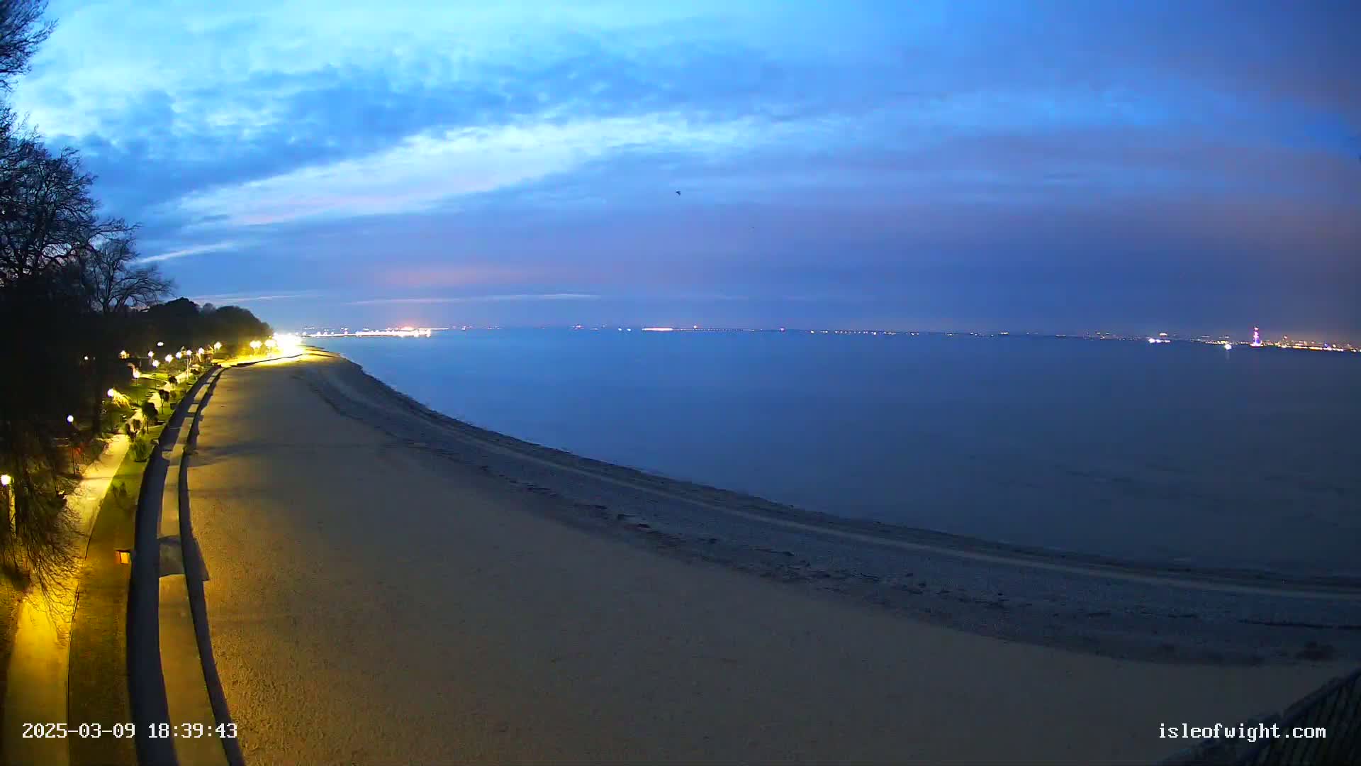 A sandy beach at twilight, with a paved walkway lined with lights leading to a calm, dark-blue sea under a mostly clear sky.
