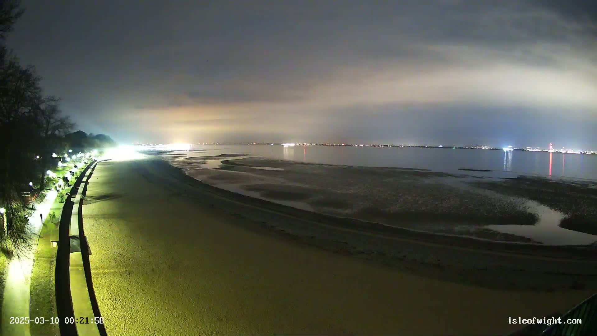 A nighttime view of a sandy beach and a paved walkway, illuminated by lights, with a calm body of water and a distant city skyline under a cloudy sky.