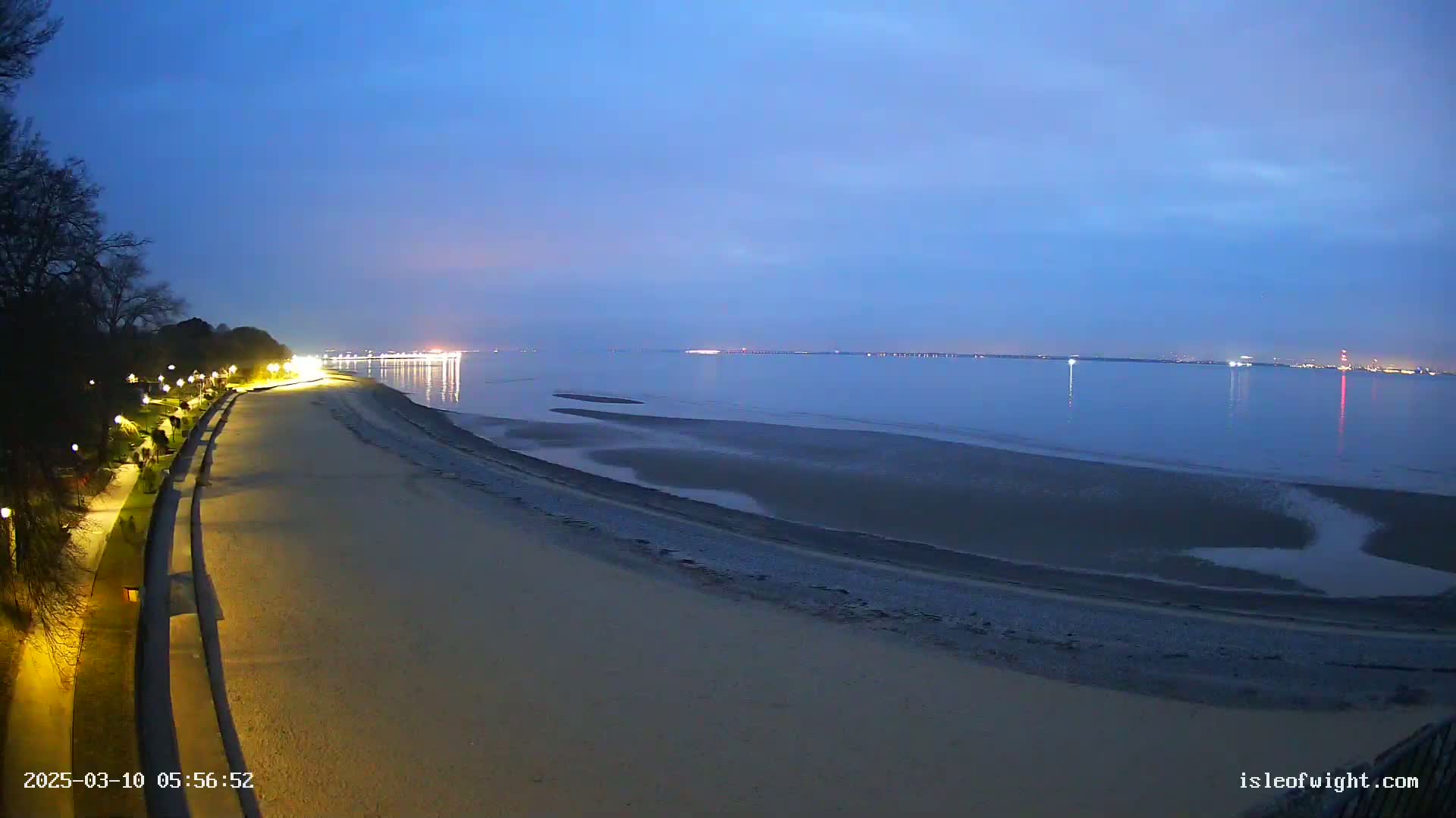 A sandy beach at twilight, illuminated by lights along a paved walkway, stretches towards a calm, dark blue body of water under a clear, dusky sky.