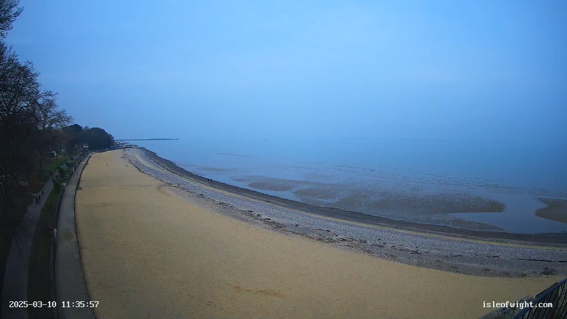 A sandy beach curves along a calm, hazy ocean under a clear blue sky.